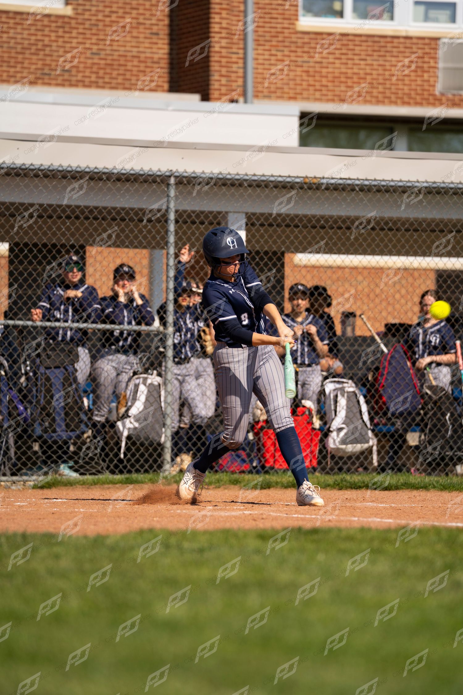 Female softball player in navy pinstripe uniform makes contact with ball at home plate during a game near school building.