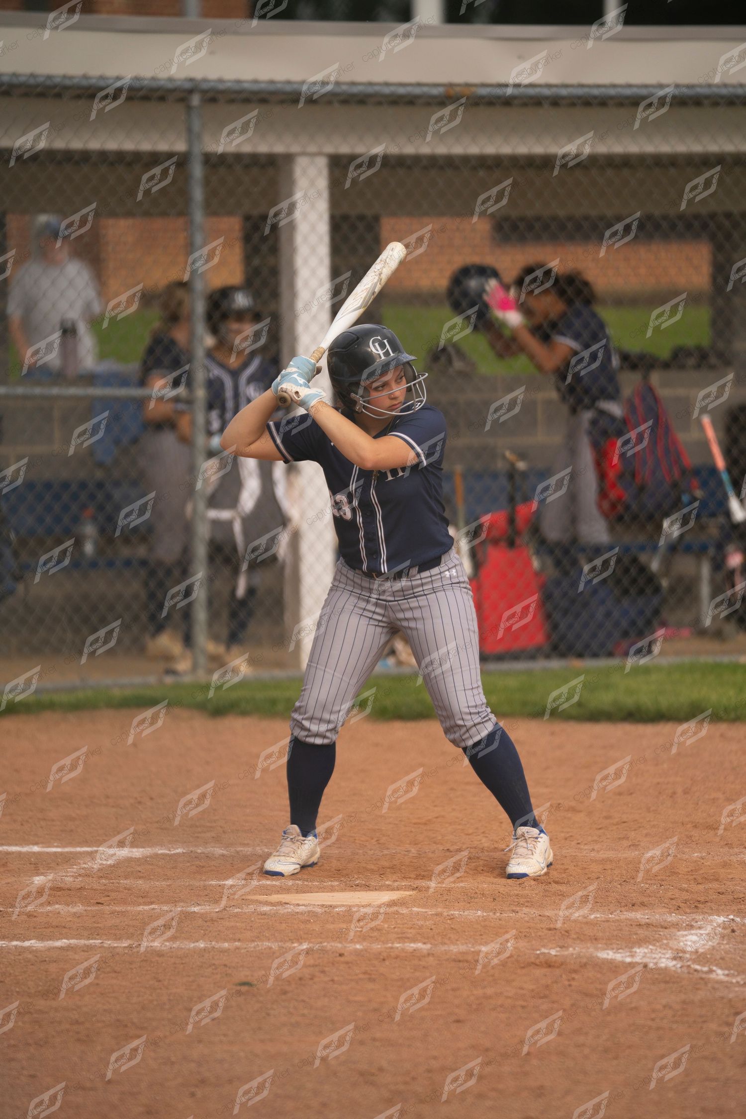 Female softball batter in navy pinstripe uniform in batting stance at home plate.