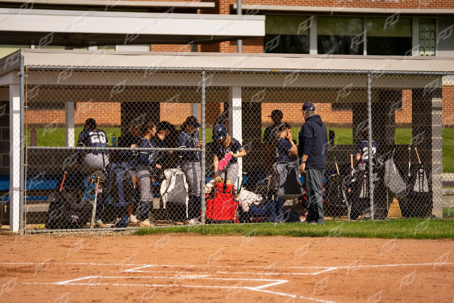 Softball team gathered in the dugout behind a chain-link fence during a game.