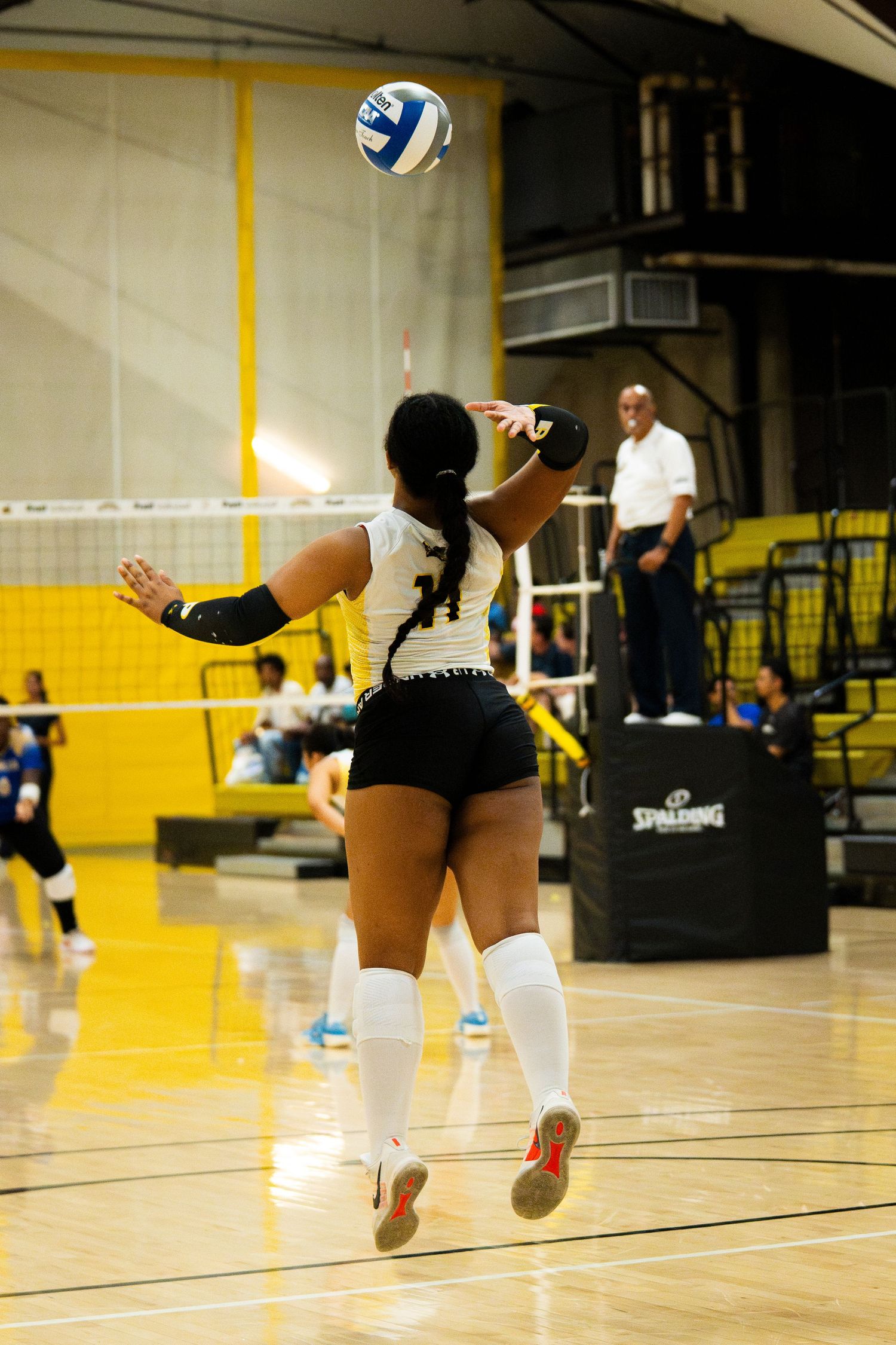 Female volleyball player in white uniform serving the ball during an indoor match on a hardwood court.