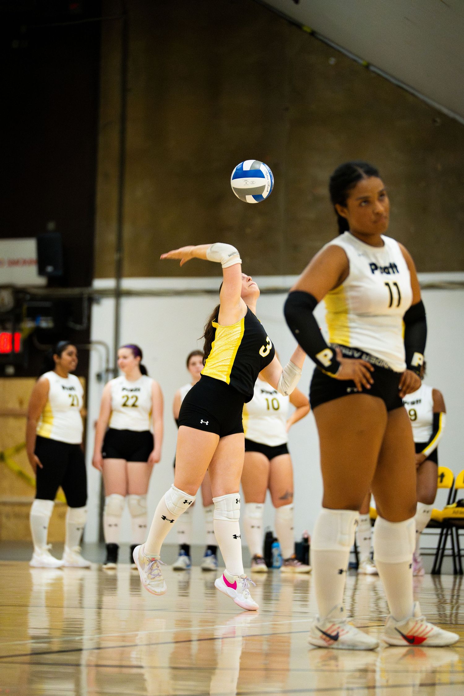 Female volleyball player serving the ball during a match, teammates in white and yellow uniforms visible in background.
