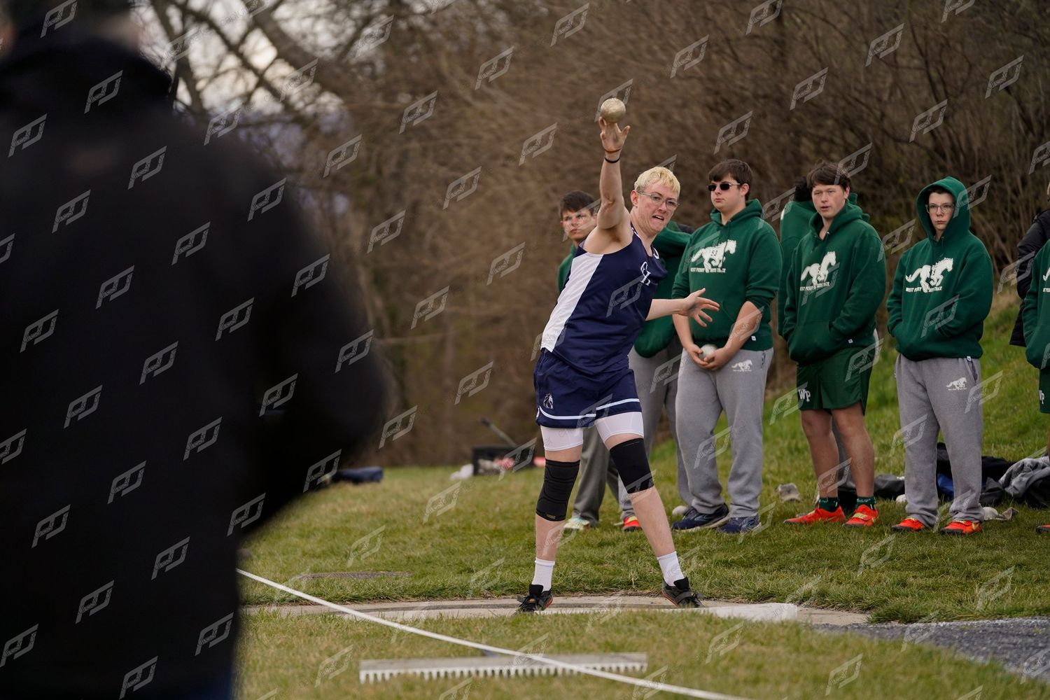 High school athlete prepares to release a shot put with teammates watching in the background.