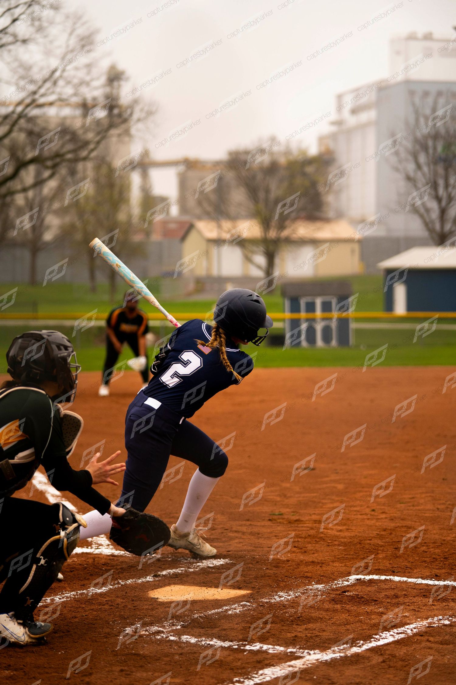Softball batter number 2 in navy uniform swings at home plate with catcher crouching behind her.