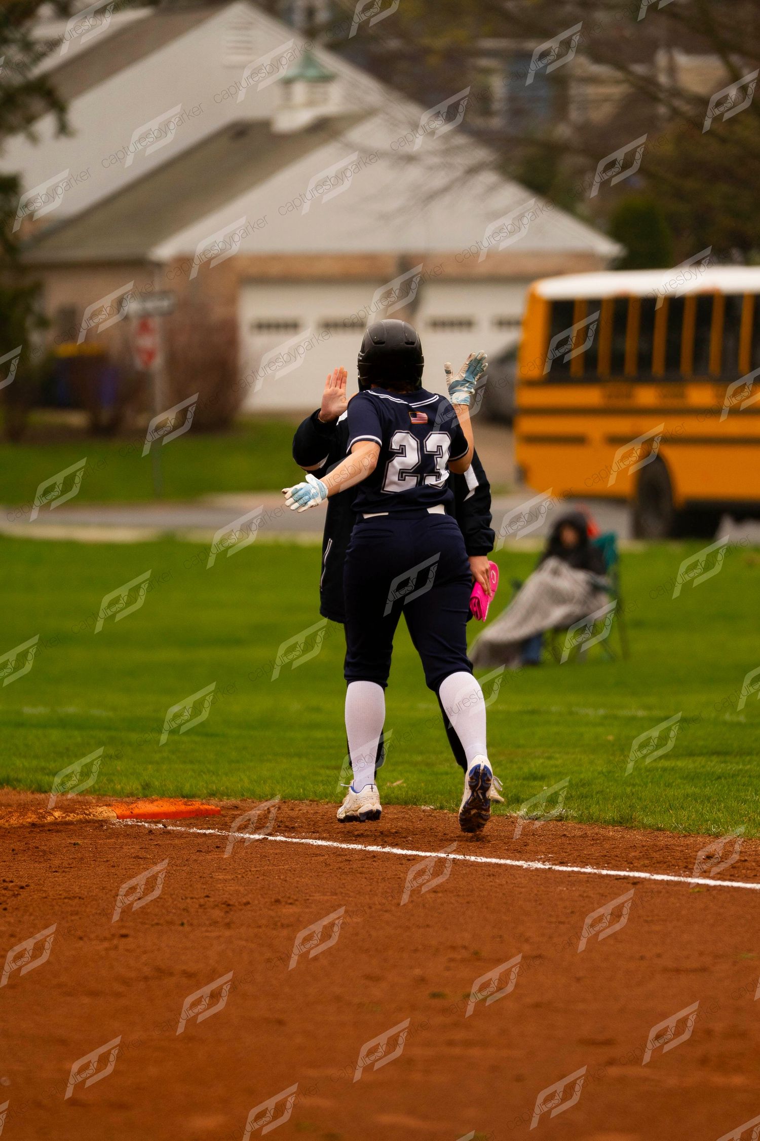 Softball player #23 in navy uniform runs bases with hand raised during outdoor game.