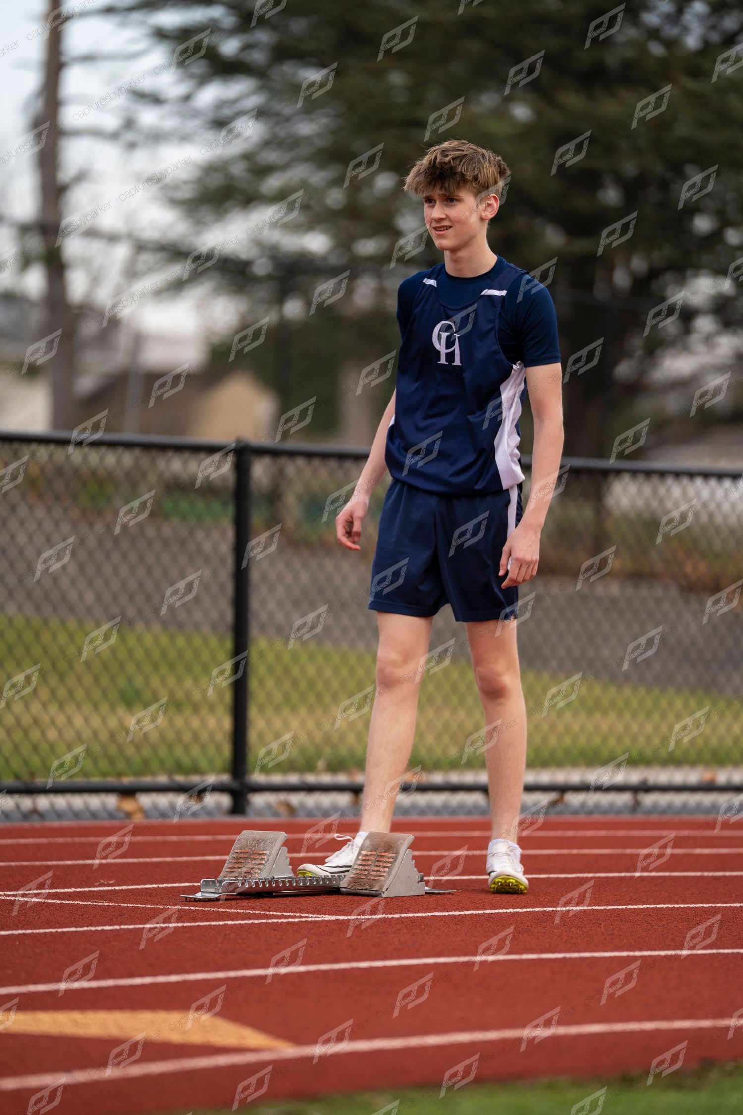 Male track athlete in navy uniform stands near starting blocks on red track.