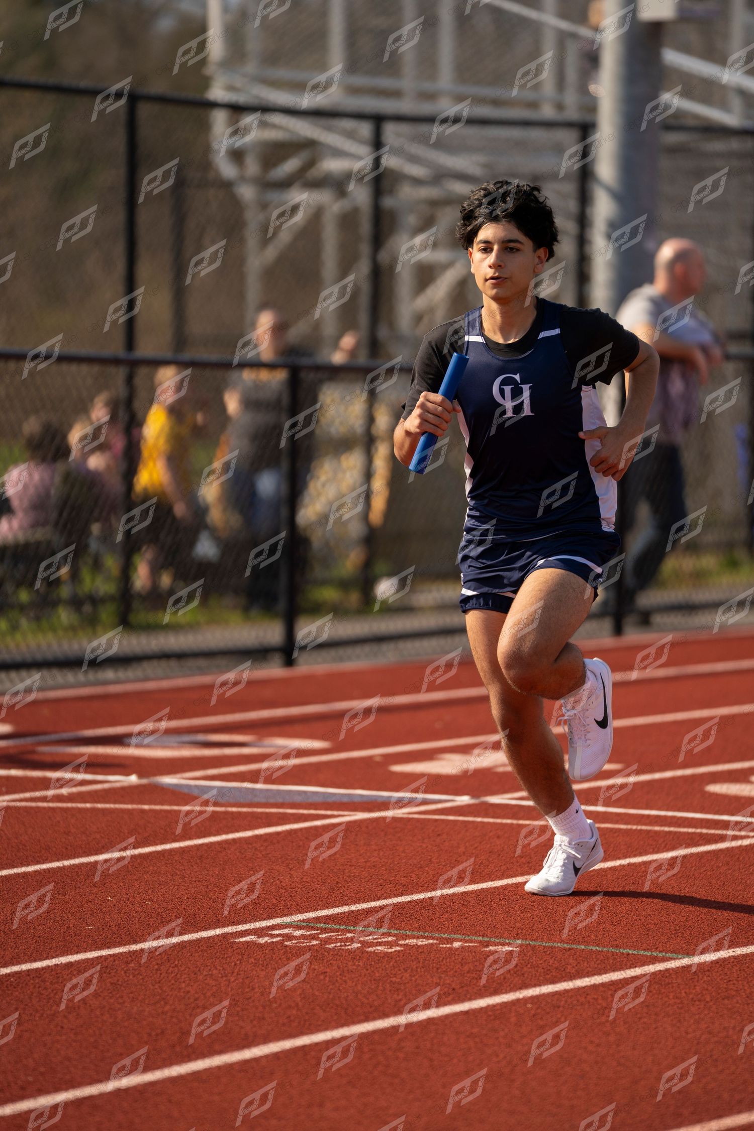 Young male track athlete in navy CH uniform runs relay race holding blue baton on red track.