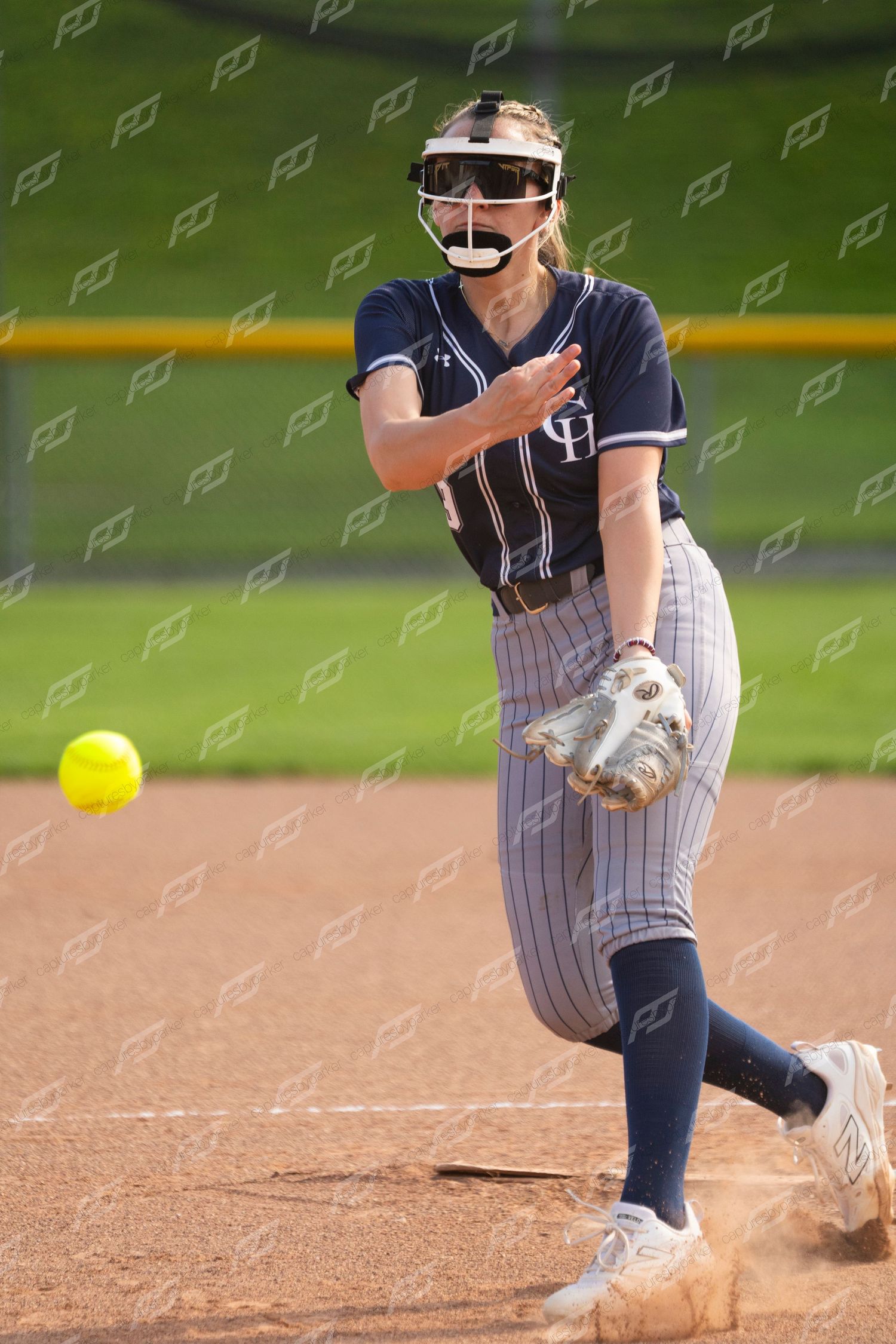 Female softball pitcher in navy uniform releases a yellow ball during a game.