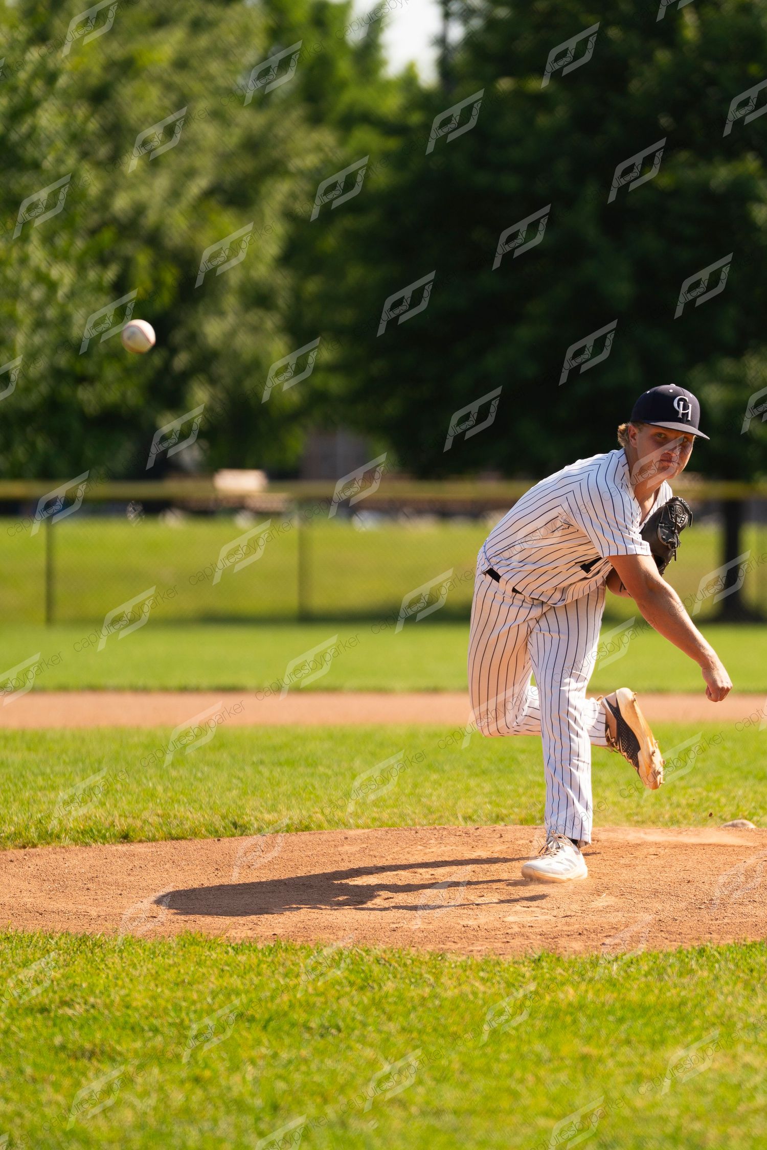 Youth baseball pitcher in white pinstripe uniform releasing a pitch from the mound on a sunny day.