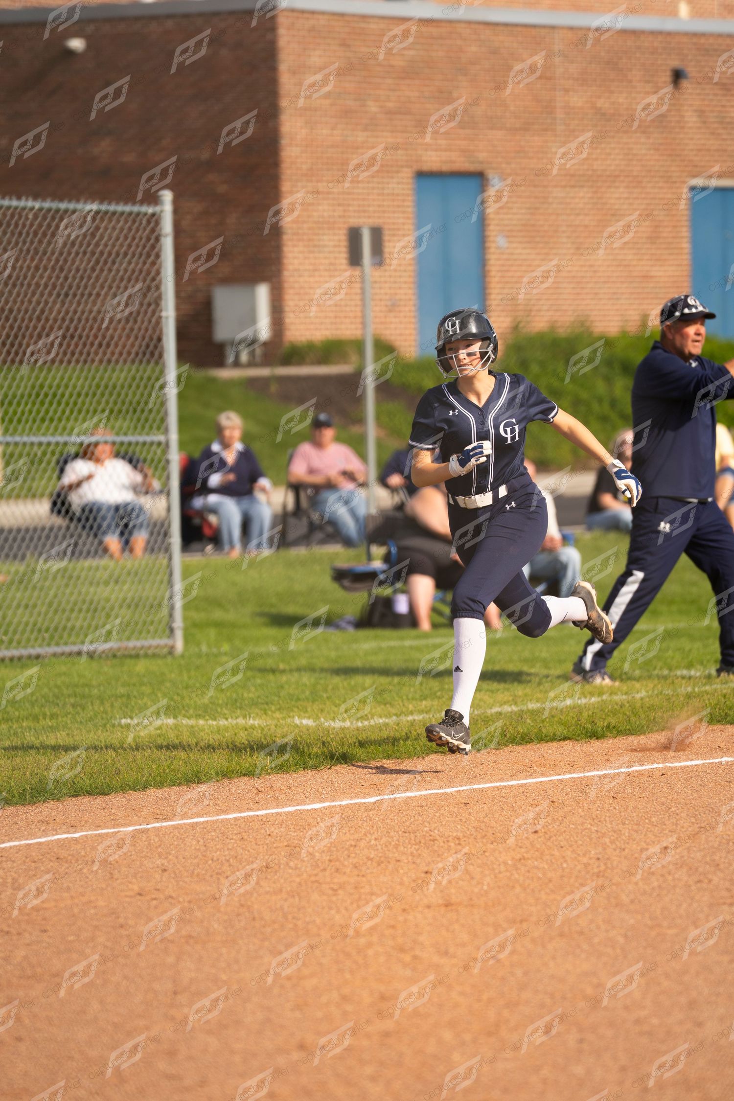 A softball player in navy uniform sprints along the baseline during an evening game with spectators watching.