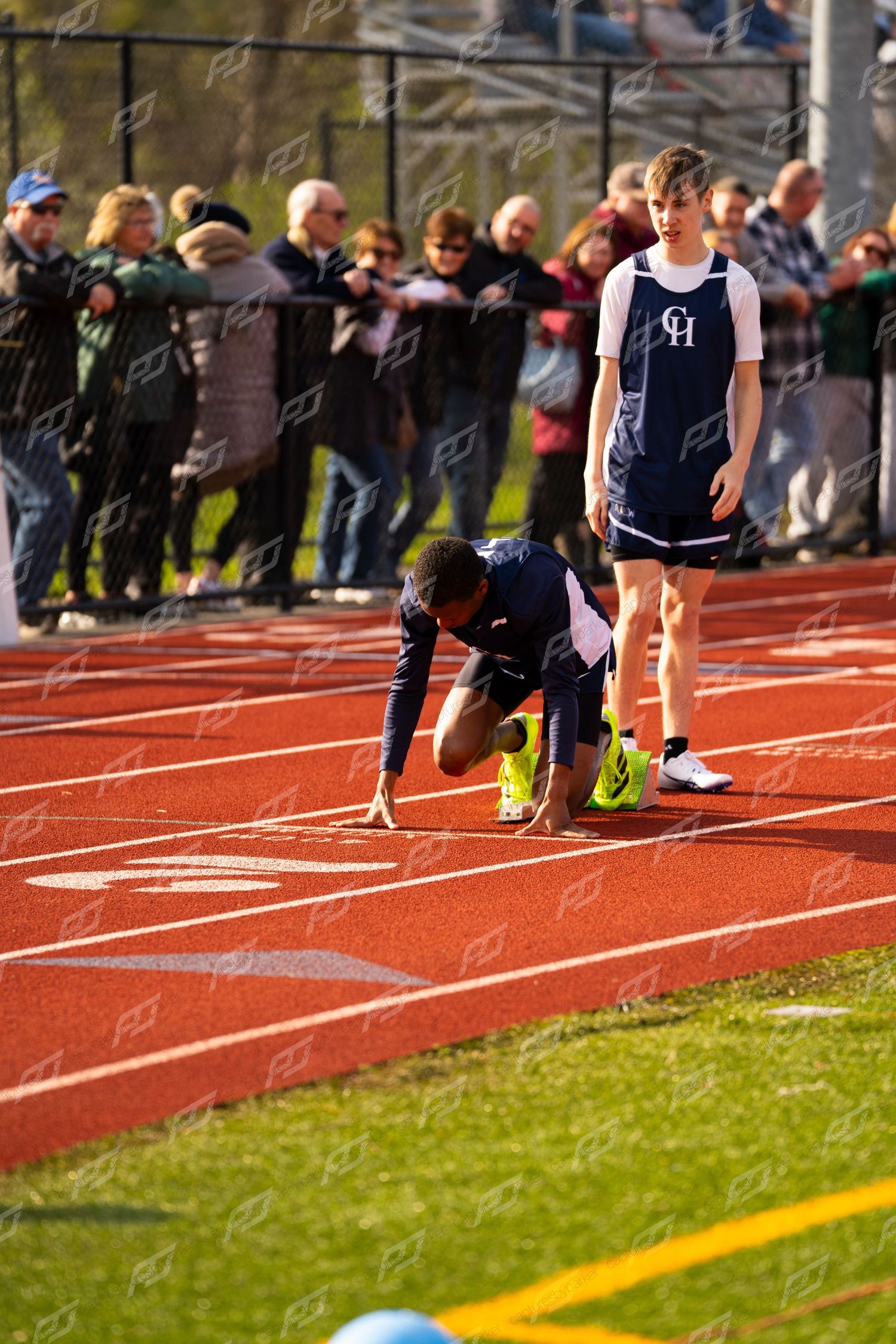 Athlete in starting position on red track at high school meet with spectators watching.