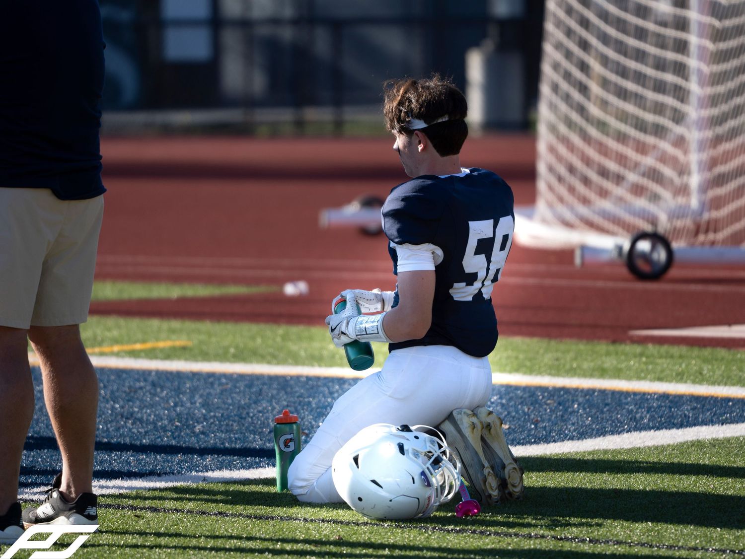 Youth football player number 58 in navy uniform kneels on turf field beside helmet during game.