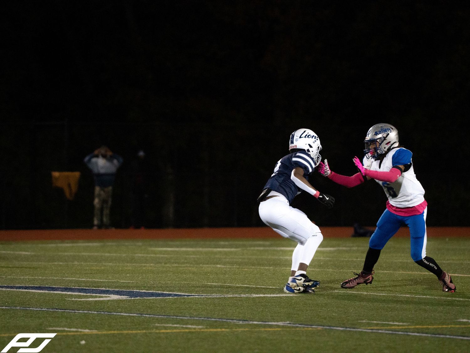 Two football players face off on dark field during high school night game under stadium lights.