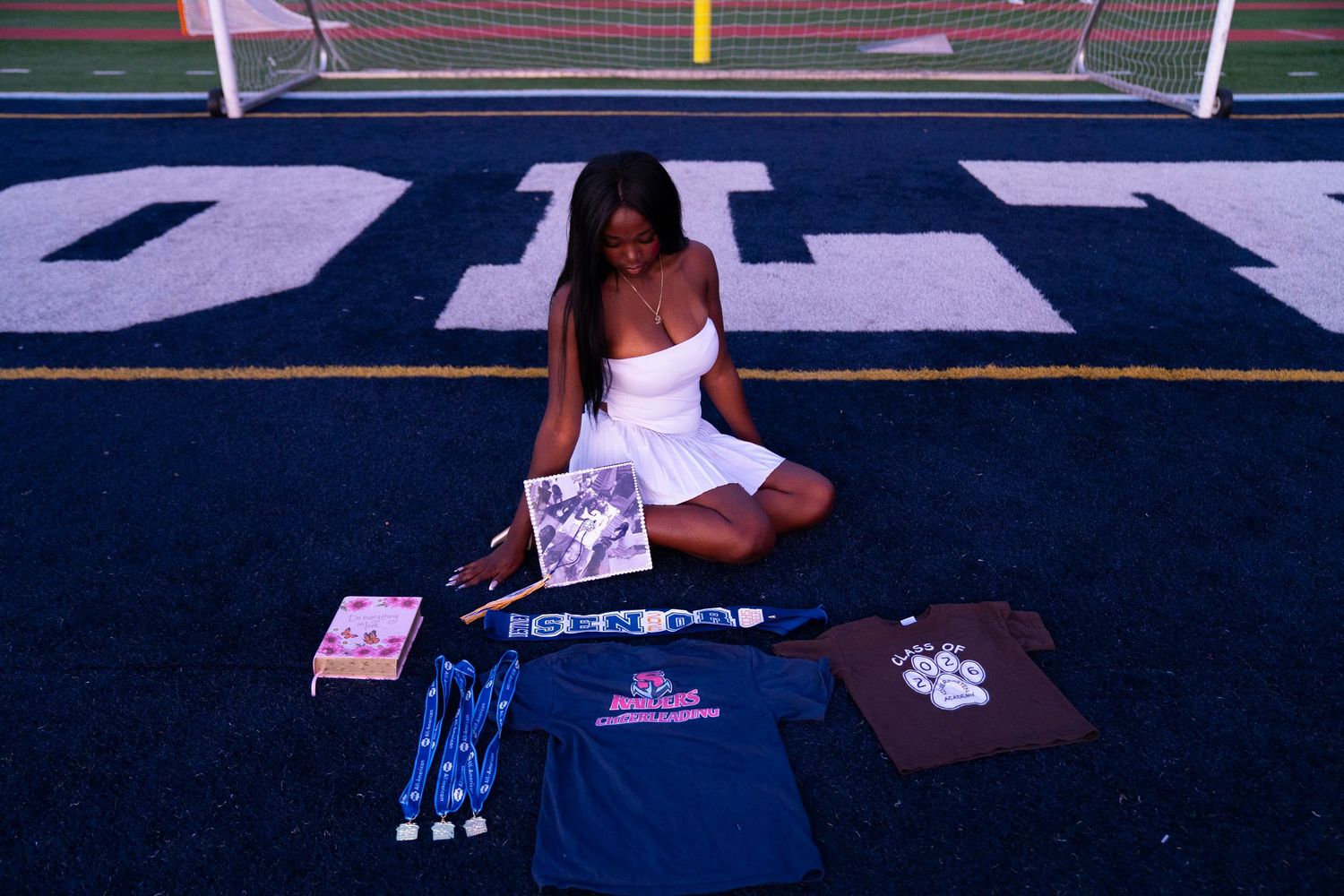 Young woman in white dress sits on turf field surrounded by college merchandise and apparel.