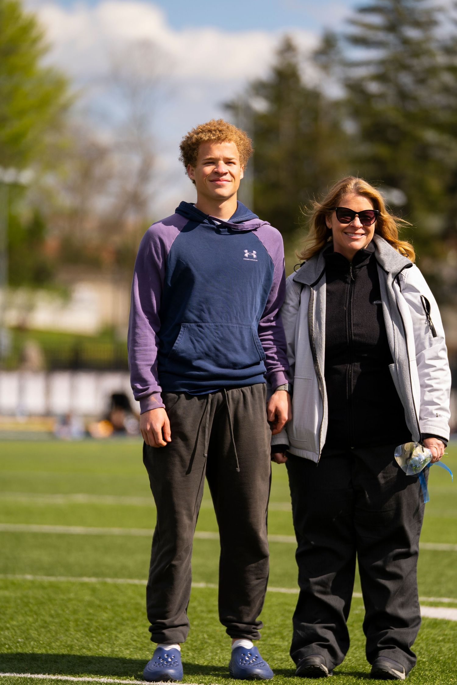 A young male athlete and a woman pose together on a sunny football field.