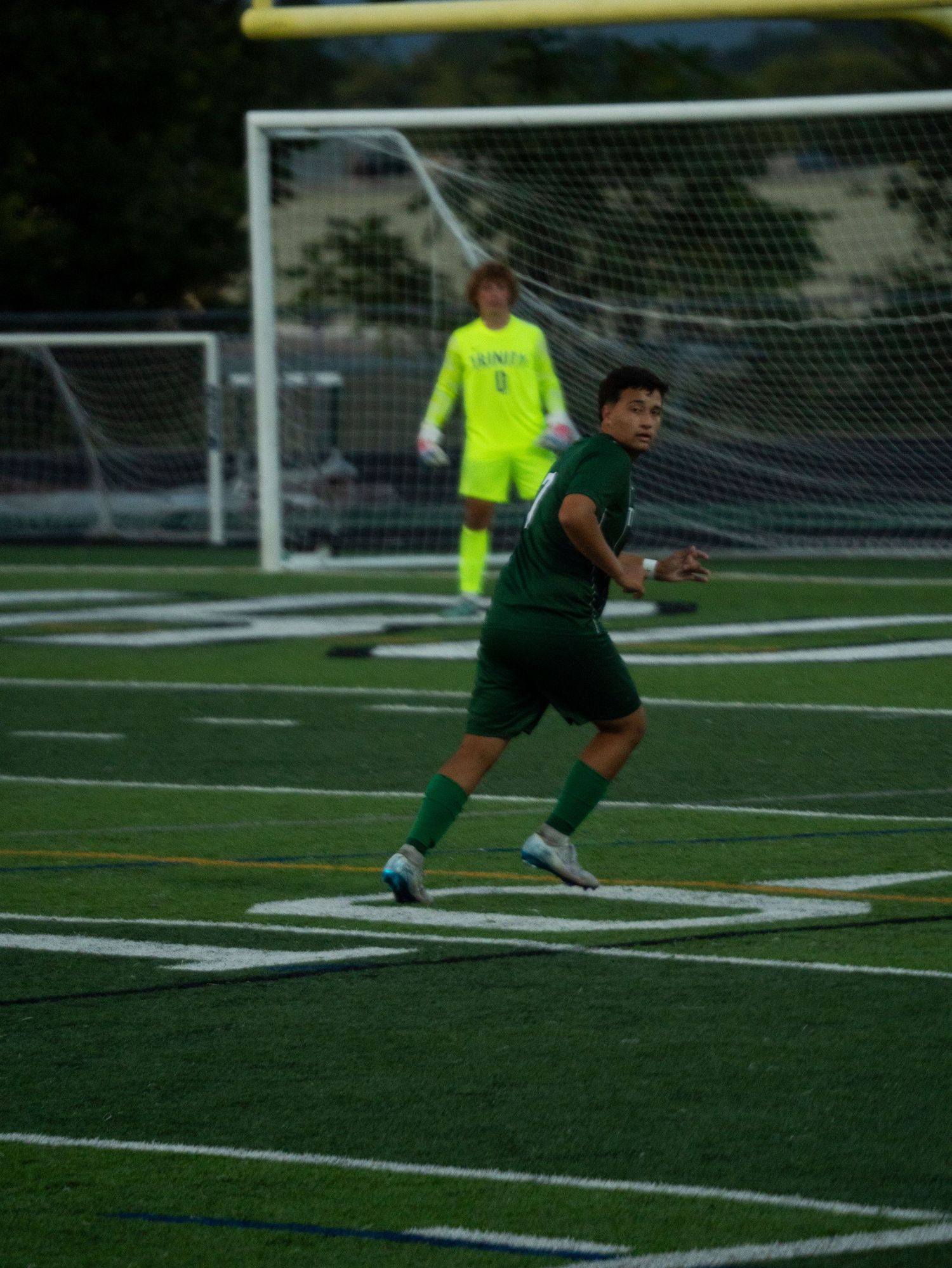 Trinity soccer player sprints on turf field with goalkeeper in yellow jersey visible near goal in background.