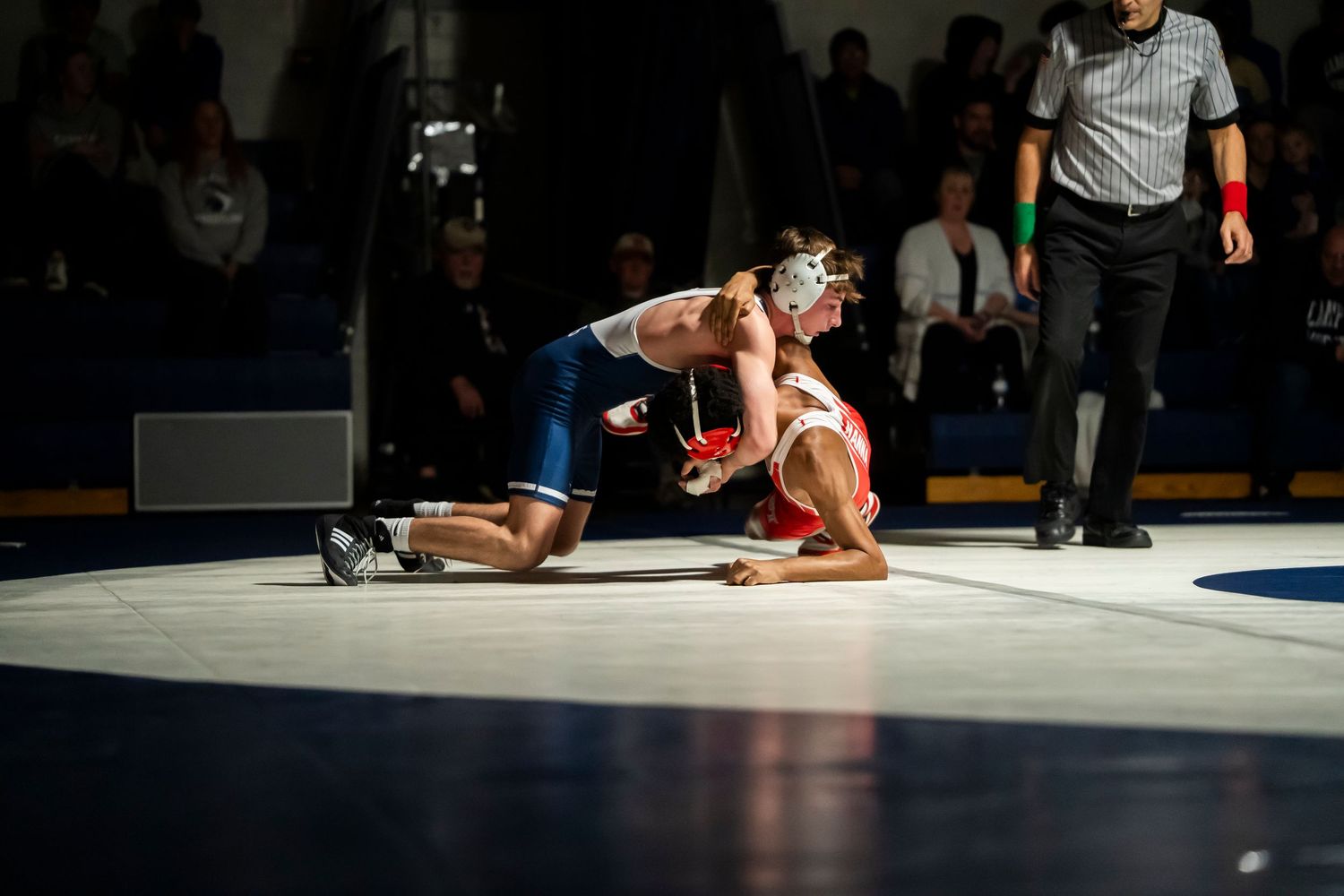 Wrestler in blue singlet controls red-clad opponent on the mat as referee watches closely.