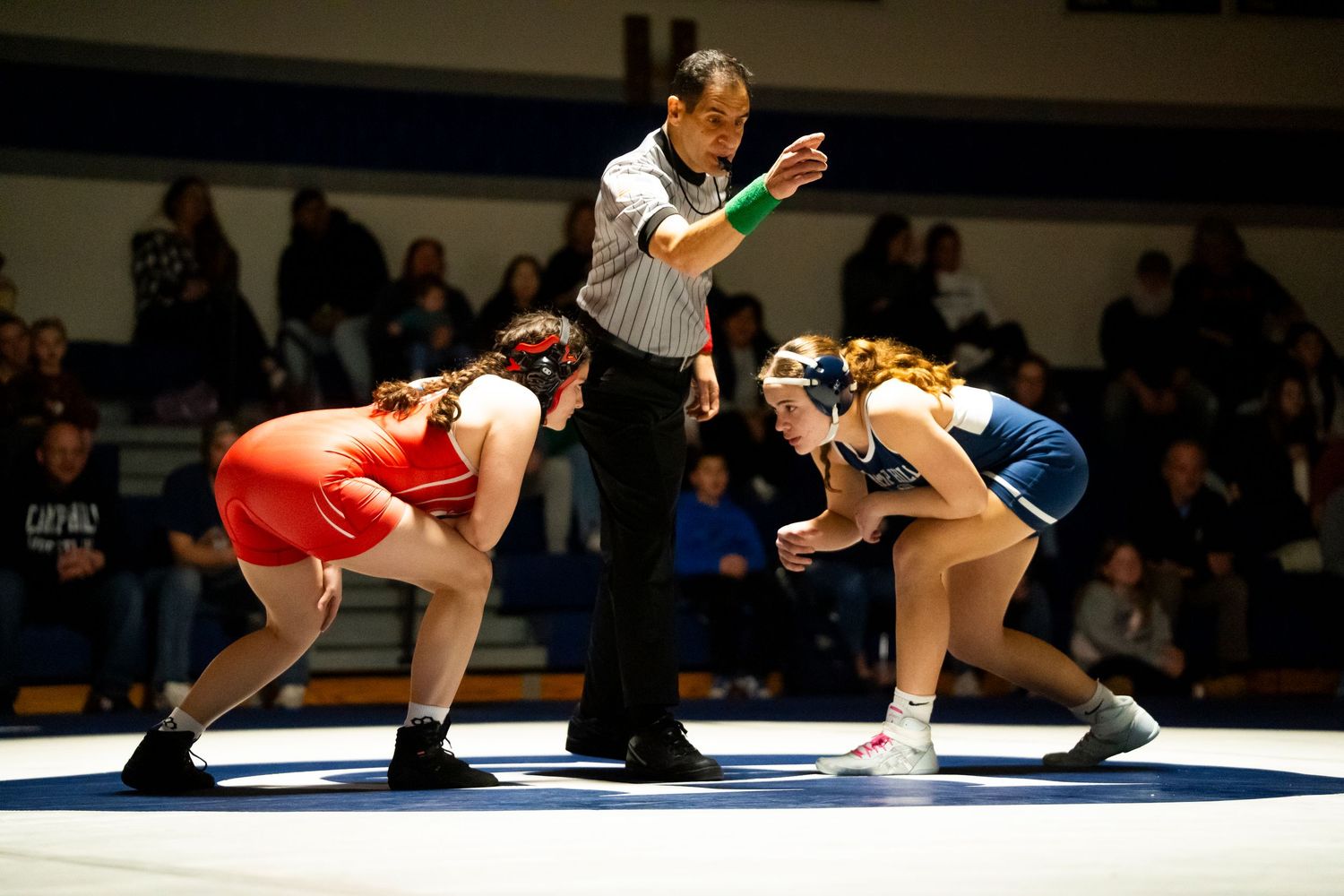 A referee signals during a girls high school wrestling match between red and blue teams.