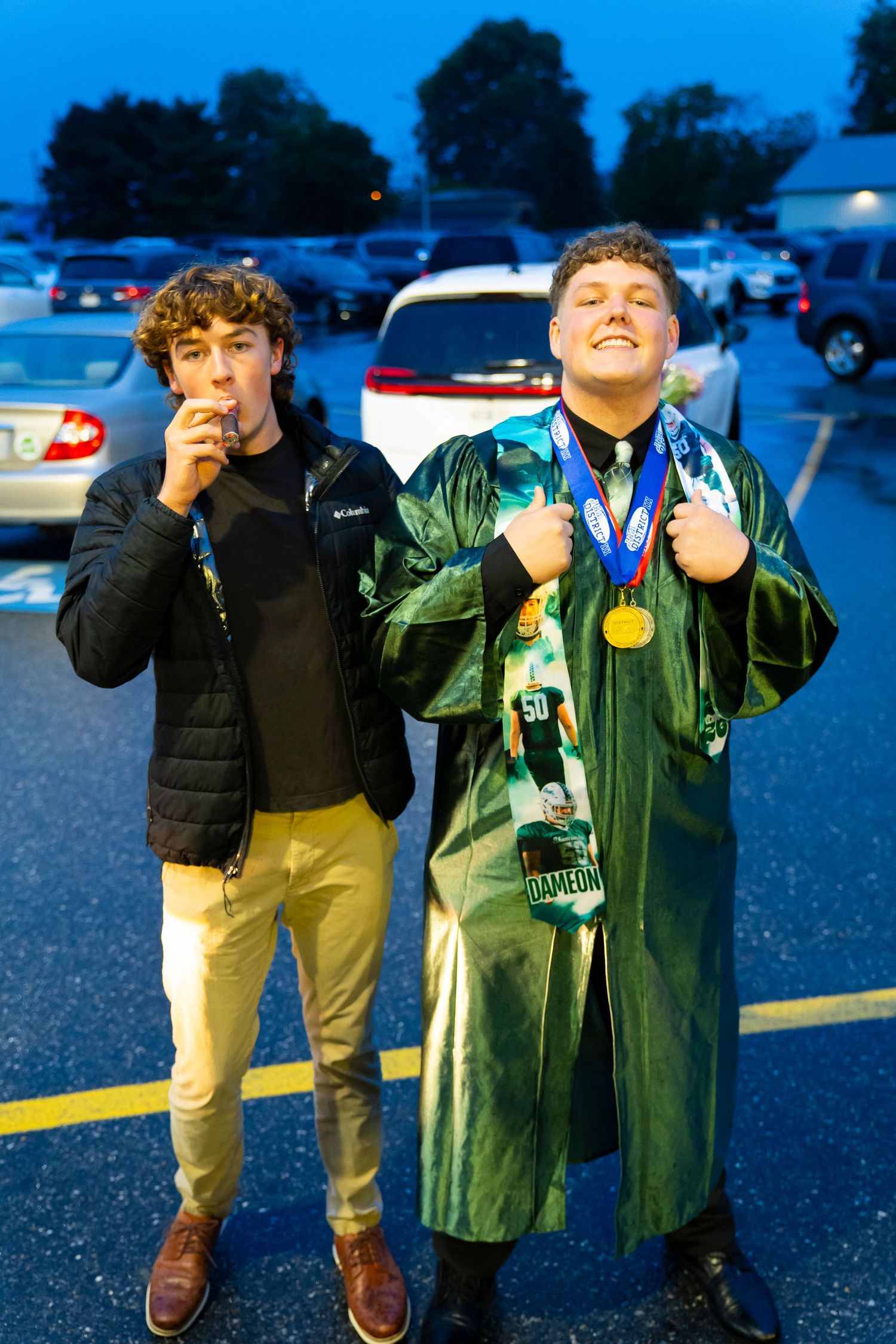 Two young men celebrate graduation, one wearing green cap and gown with gold medal in parking lot.