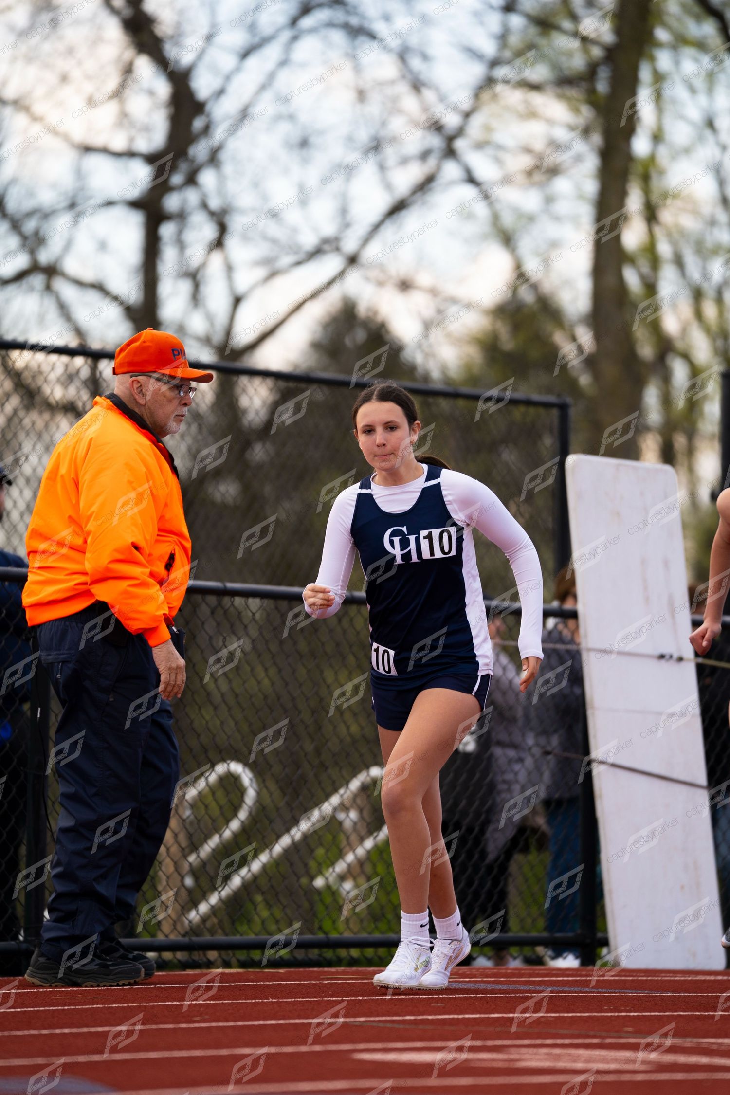 Female track athlete in navy uniform runs past a coach in an orange jacket during an outdoor track meet.
