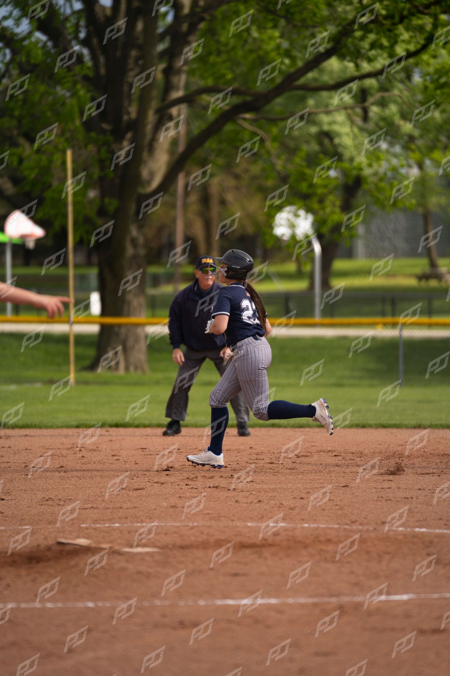 Female softball player #23 rounds a base with an umpire watching during a softball game.