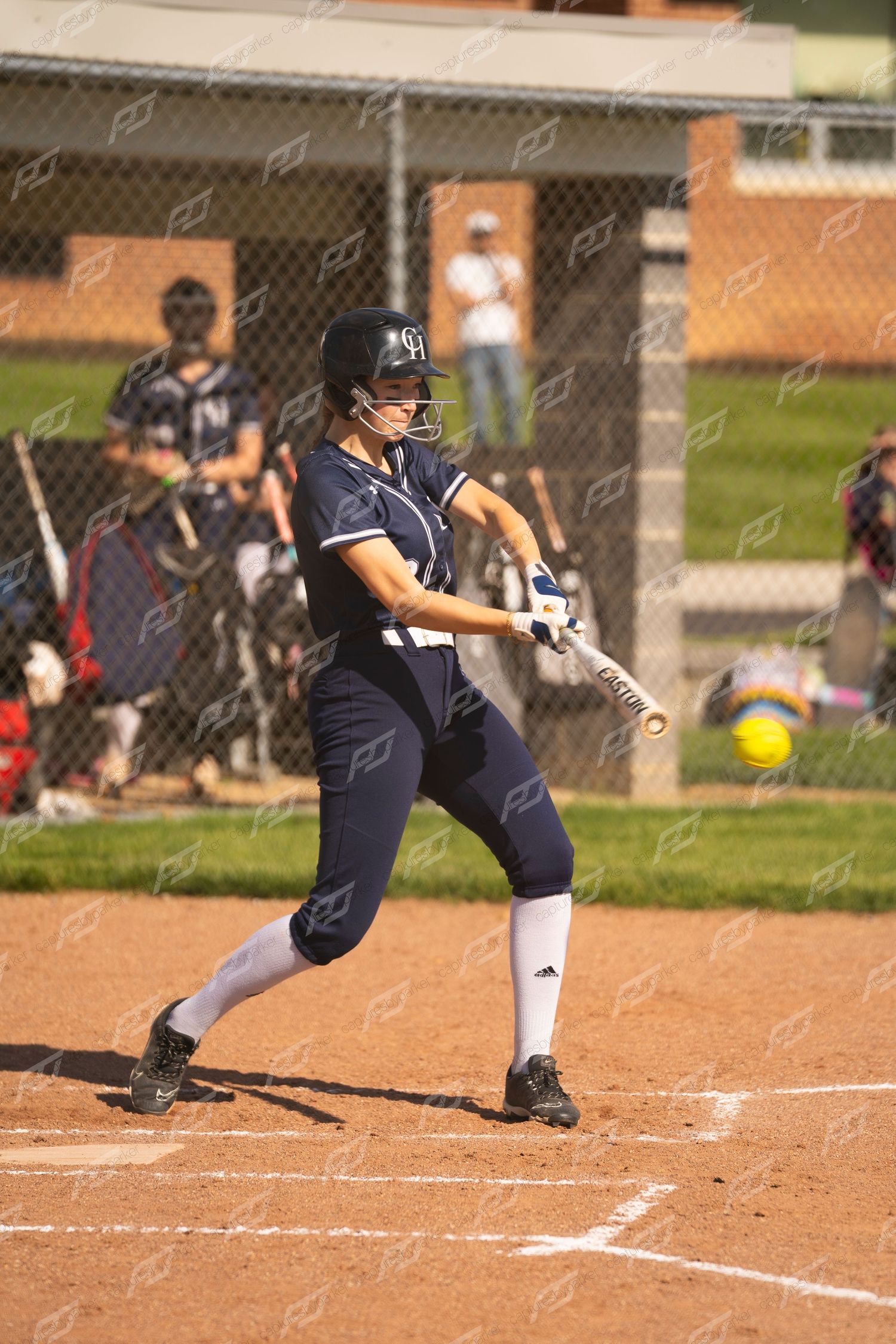 Female softball player in navy Adidas uniform making contact with the ball at home plate.