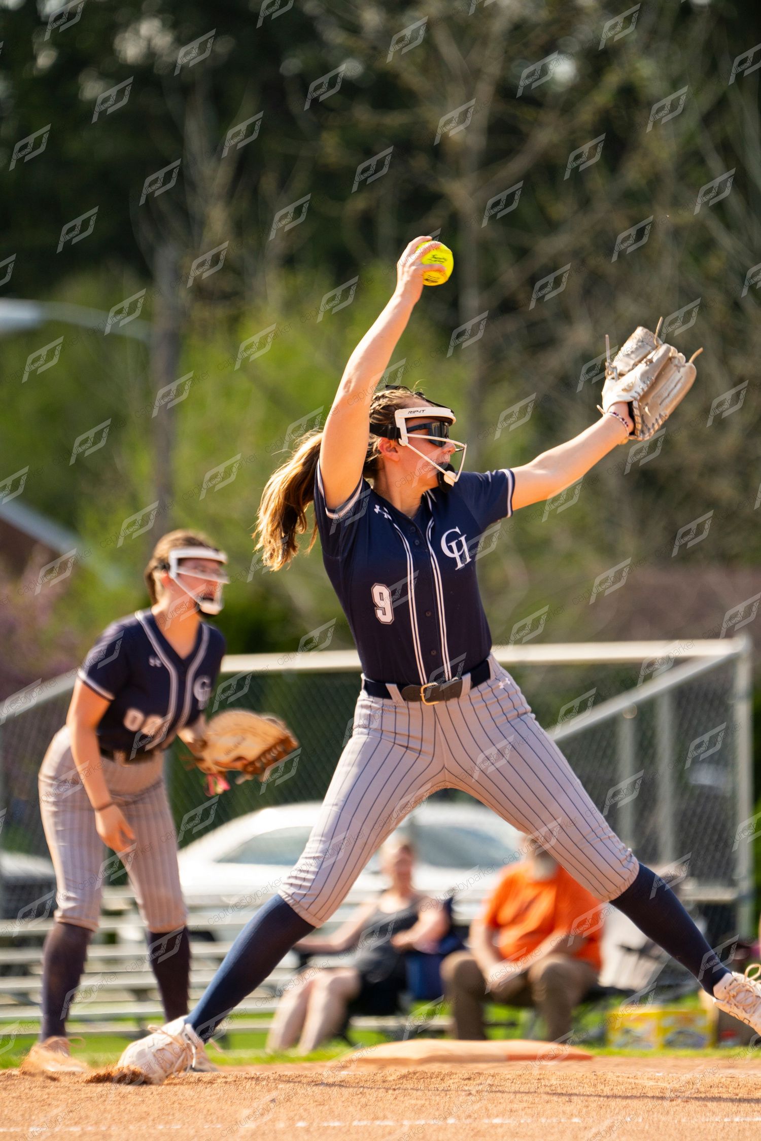 Female softball pitcher number 9 in navy uniform delivers windmill pitch during competitive game.
