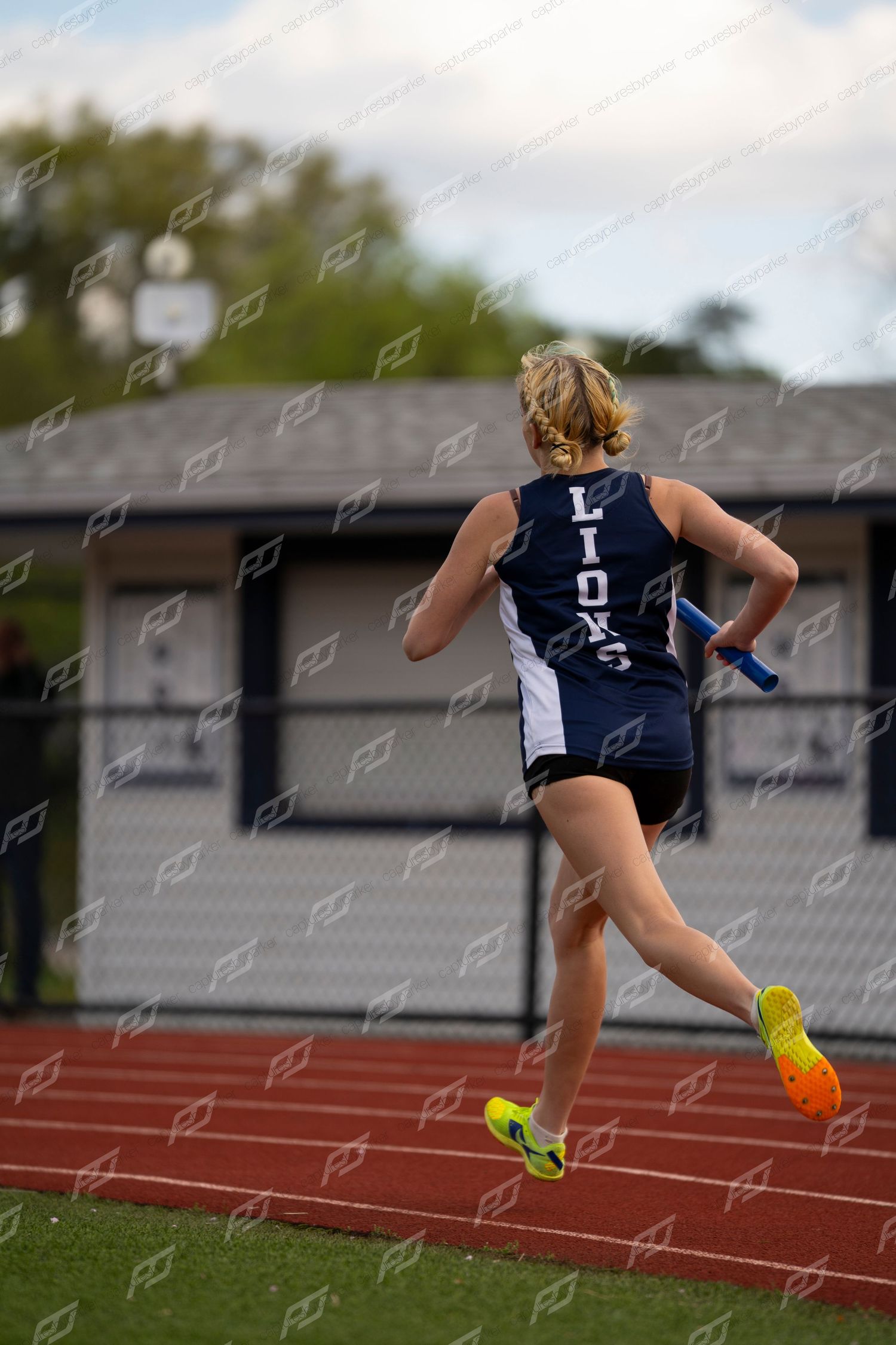 Female Lions track athlete running a relay race from behind, holding a blue baton on a red outdoor track.