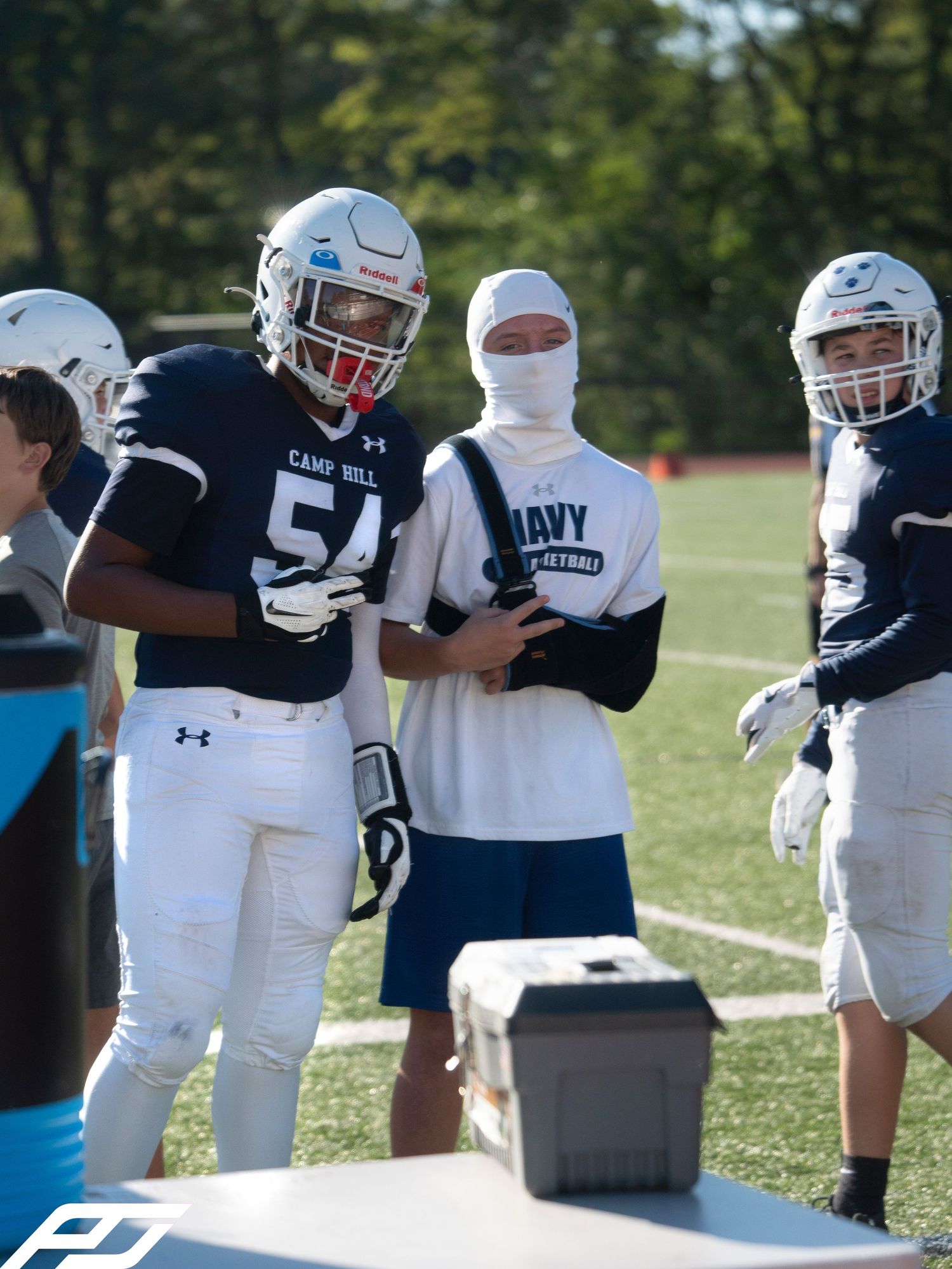 Camp Hill football player #54 stands on sideline with teammates during sunny outdoor practice.