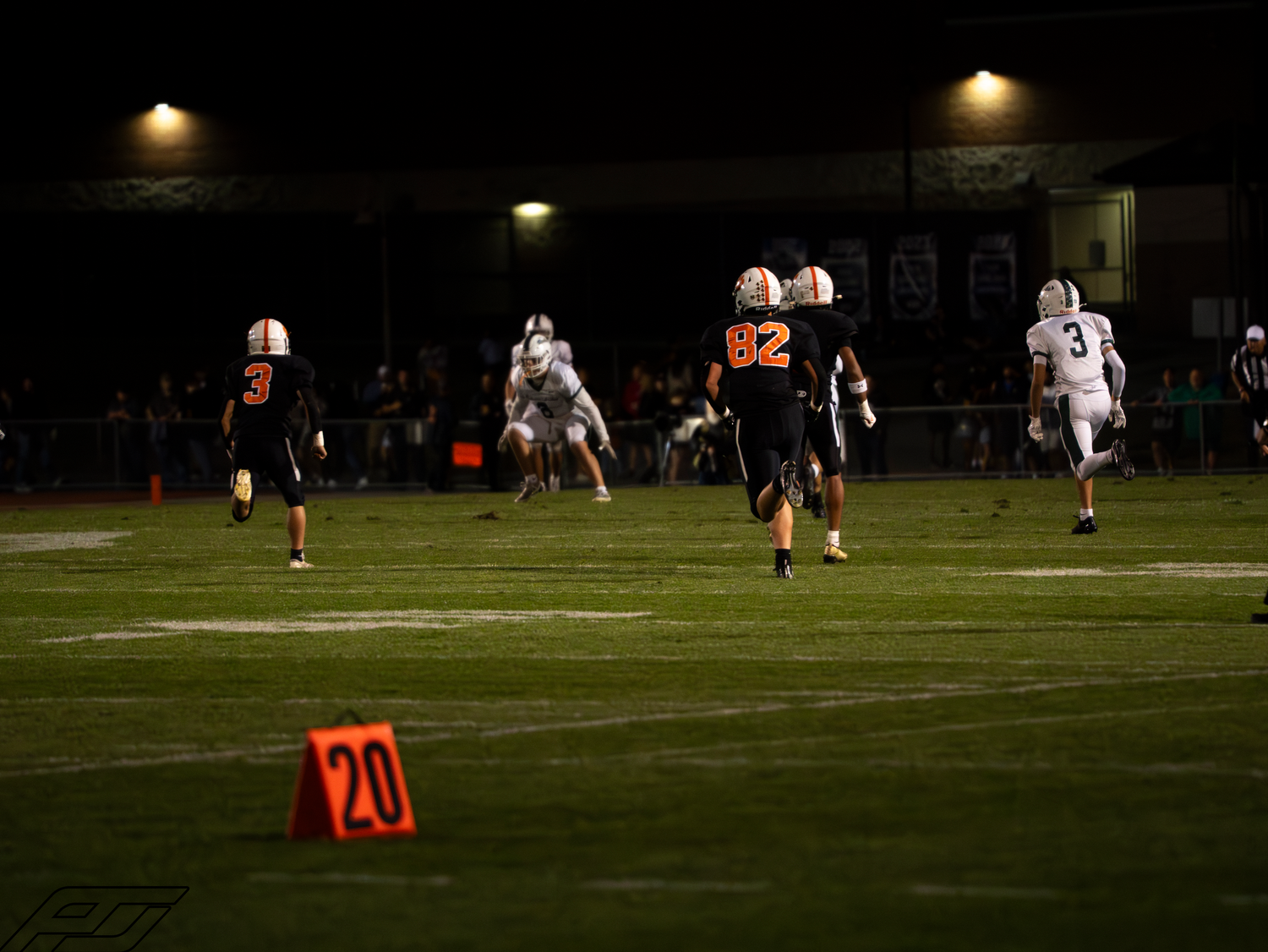 Football players in orange and white uniforms compete on a night game field near a number 20 yard marker.