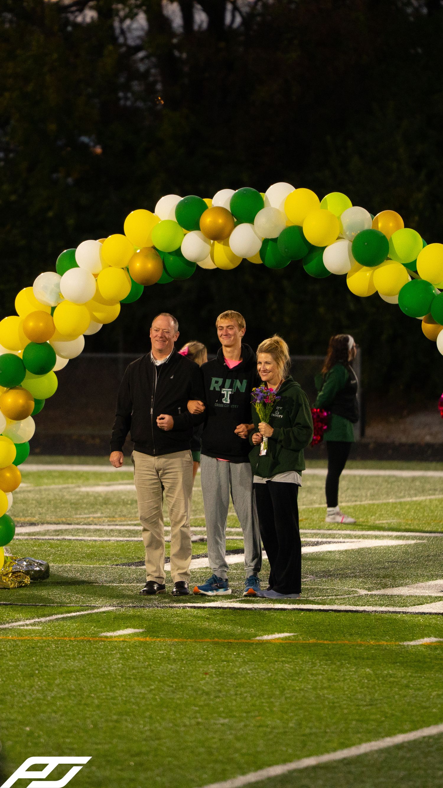 Young man in Shamrocks attire walks with parents under green and yellow balloon arch at senior night event.