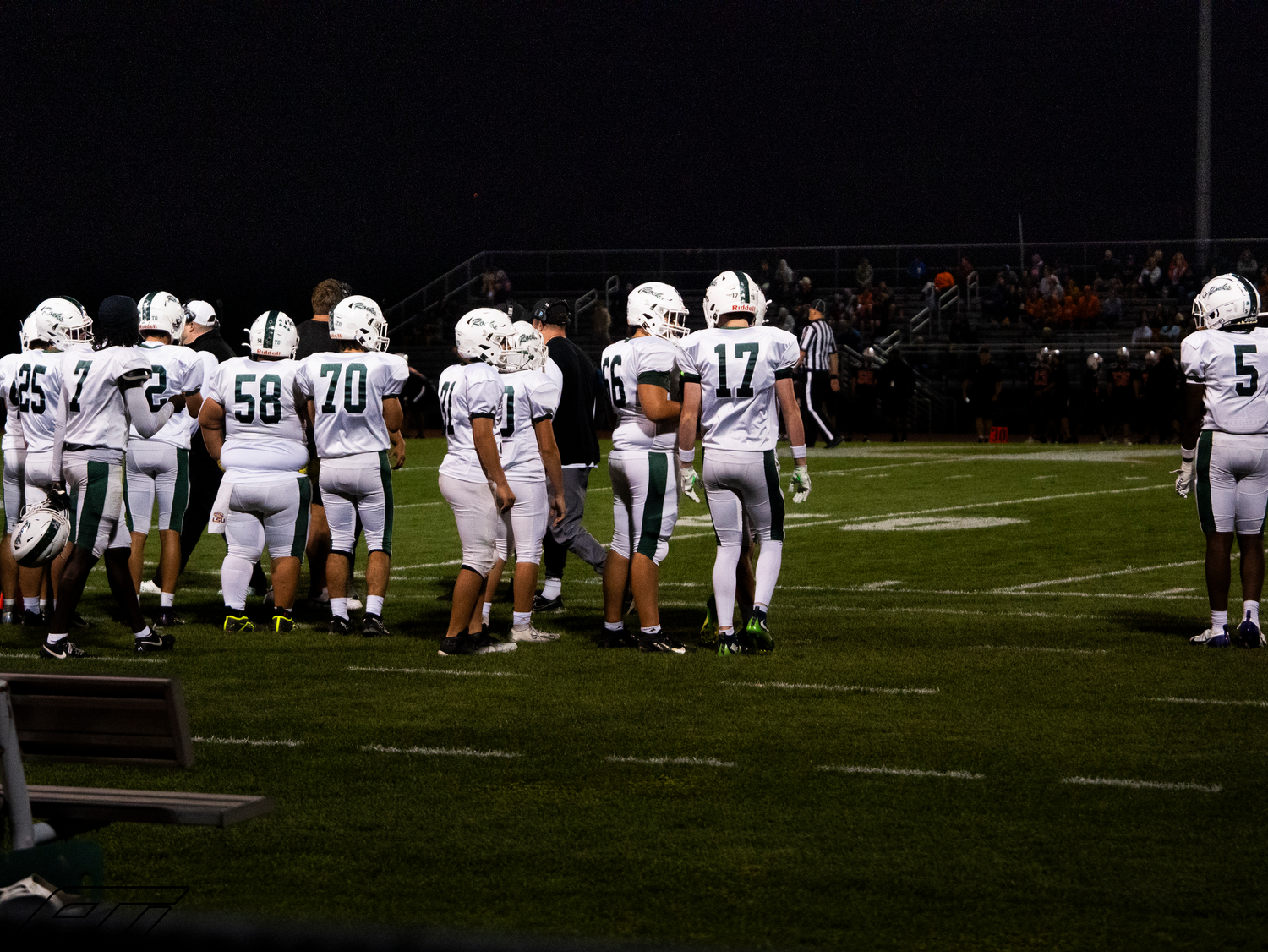 Football players in white uniforms gather on sideline during night game.
