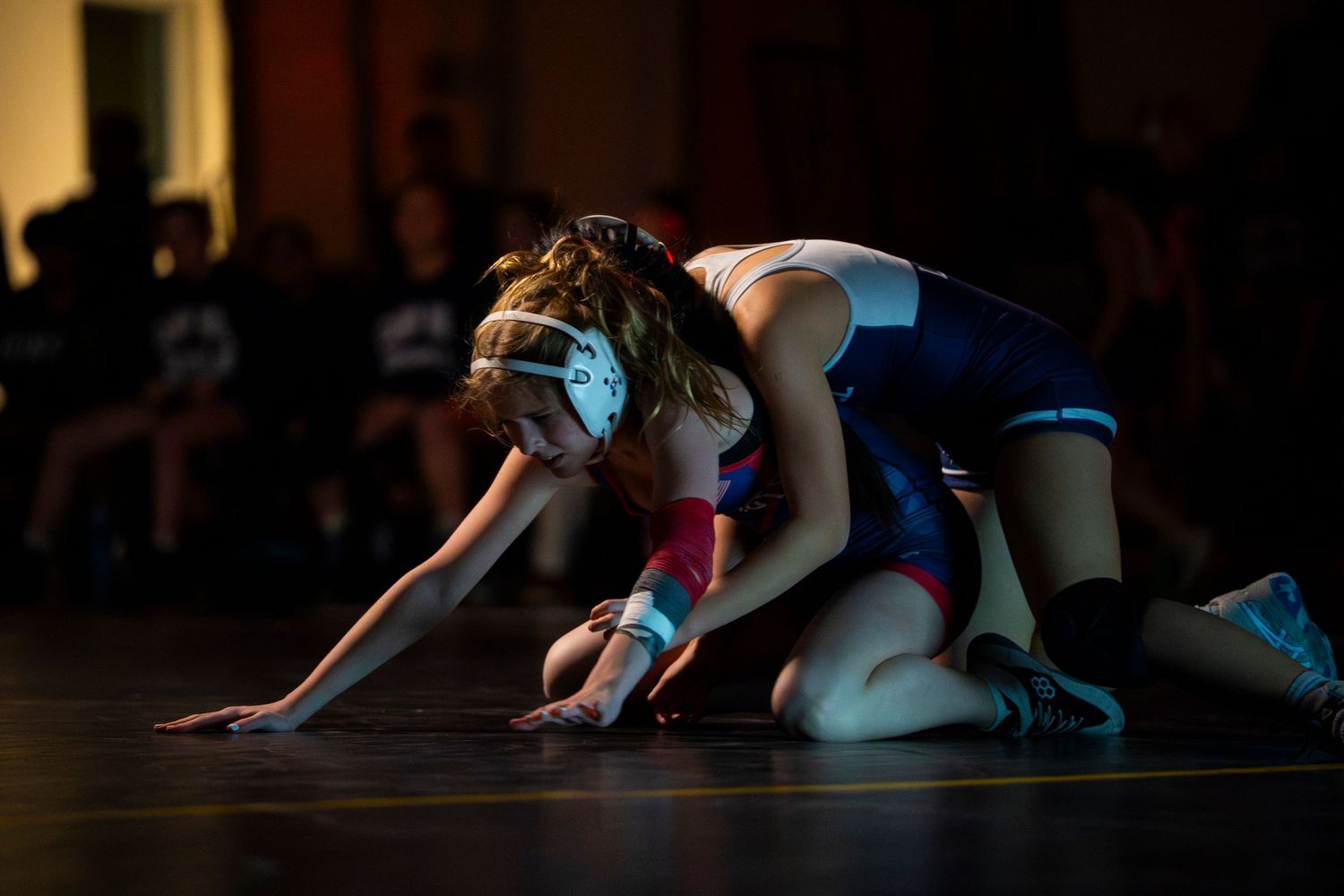 Young female wrestler in blue singlet controls opponent on dimly lit wrestling mat.