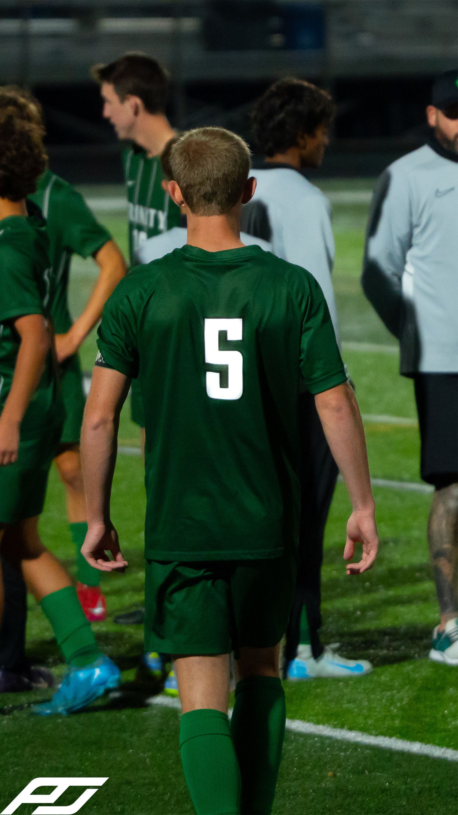 High school soccer player wearing green jersey number 5 walks on the field during a nighttime game.