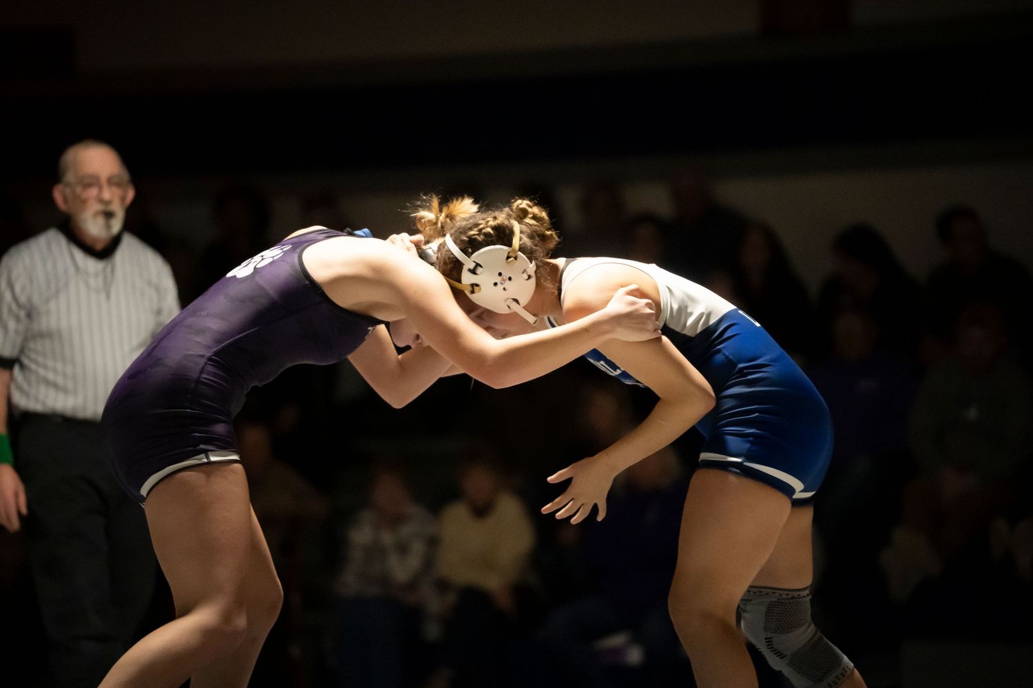 Two female wrestlers in purple and blue singlets lock up during a competitive high school wrestling match.