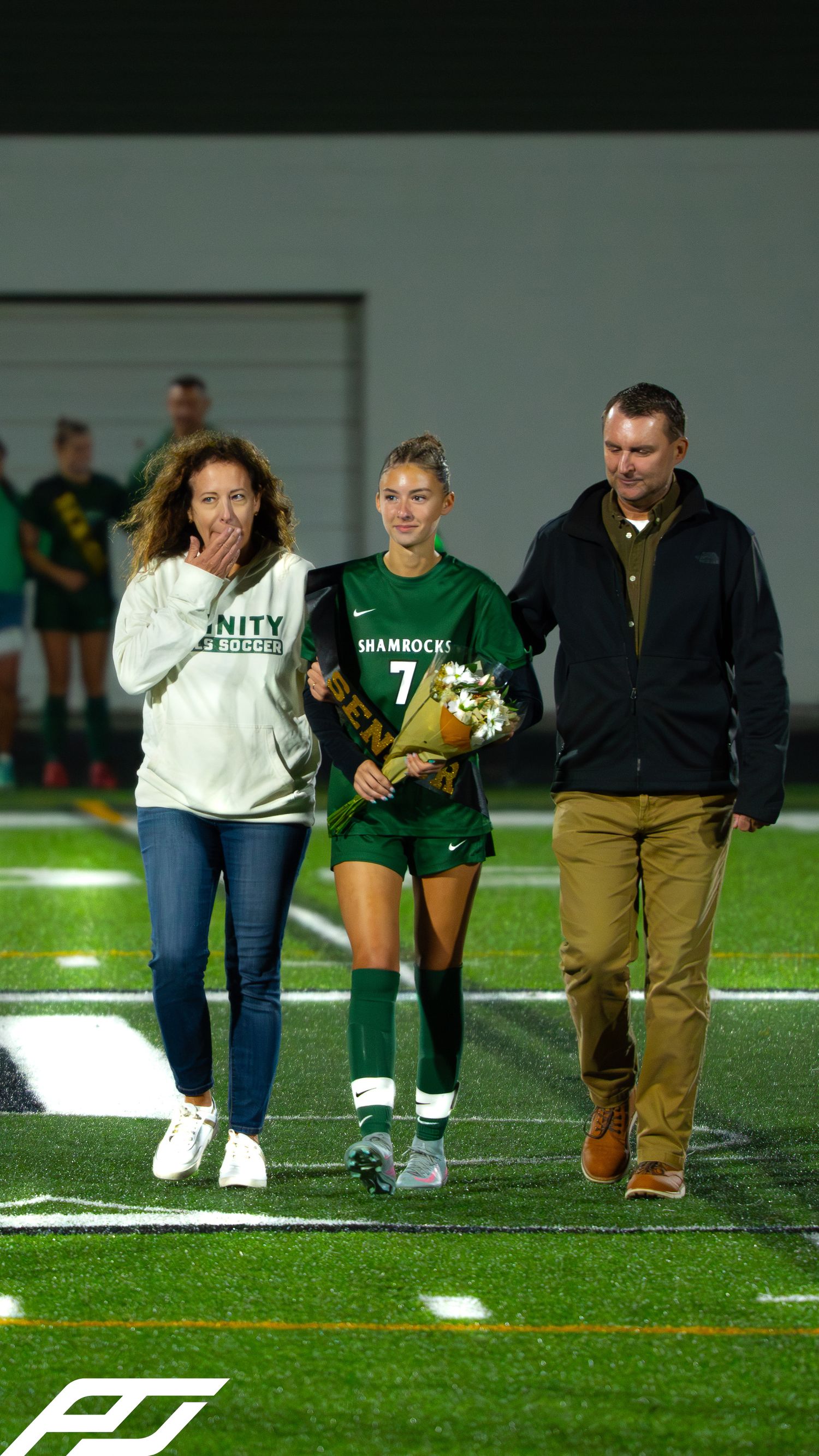 Trinity Shamrocks soccer player number 7 walks with parents during senior night on a lit football field.