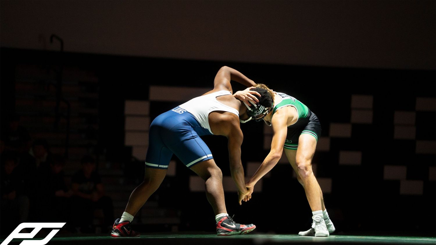 Two wrestlers compete in a grappling match under dramatic stage lighting on a gymnasium floor.