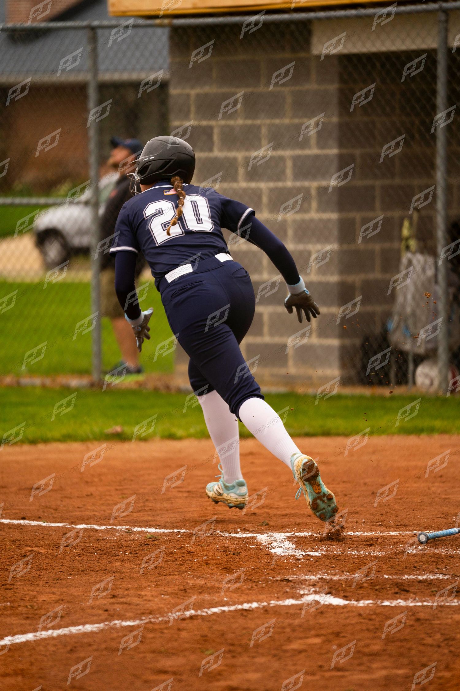 Softball player number 20 in navy uniform sprints away from home plate after hitting during a game.