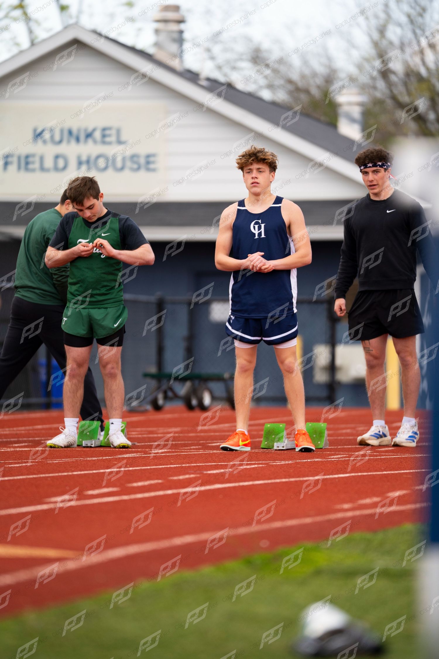 Male high school sprinter in blue GH uniform stands ready before race at outdoor track meet.