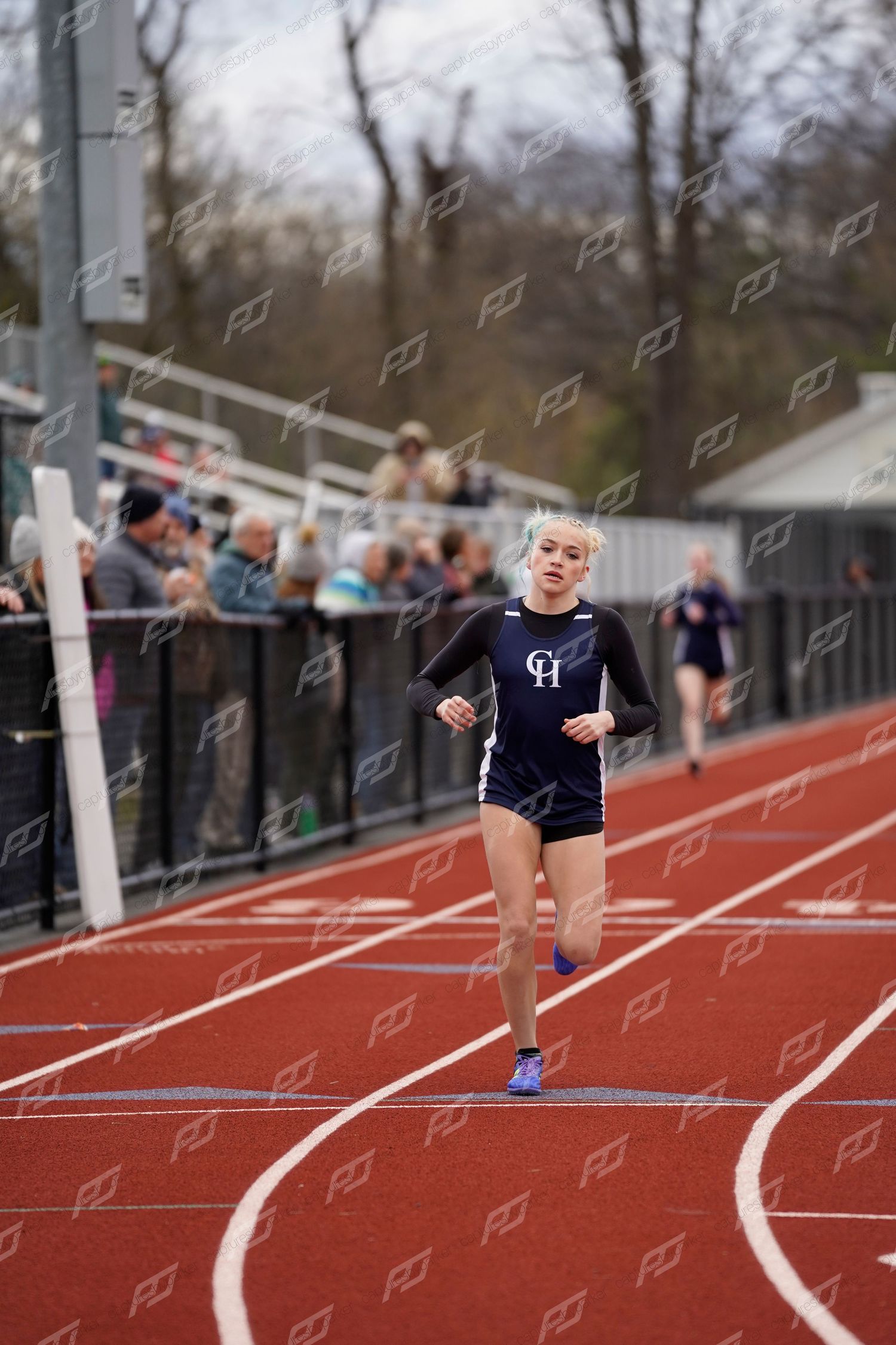 Female track athlete in navy CH uniform runs along red outdoor track during a competitive meet.