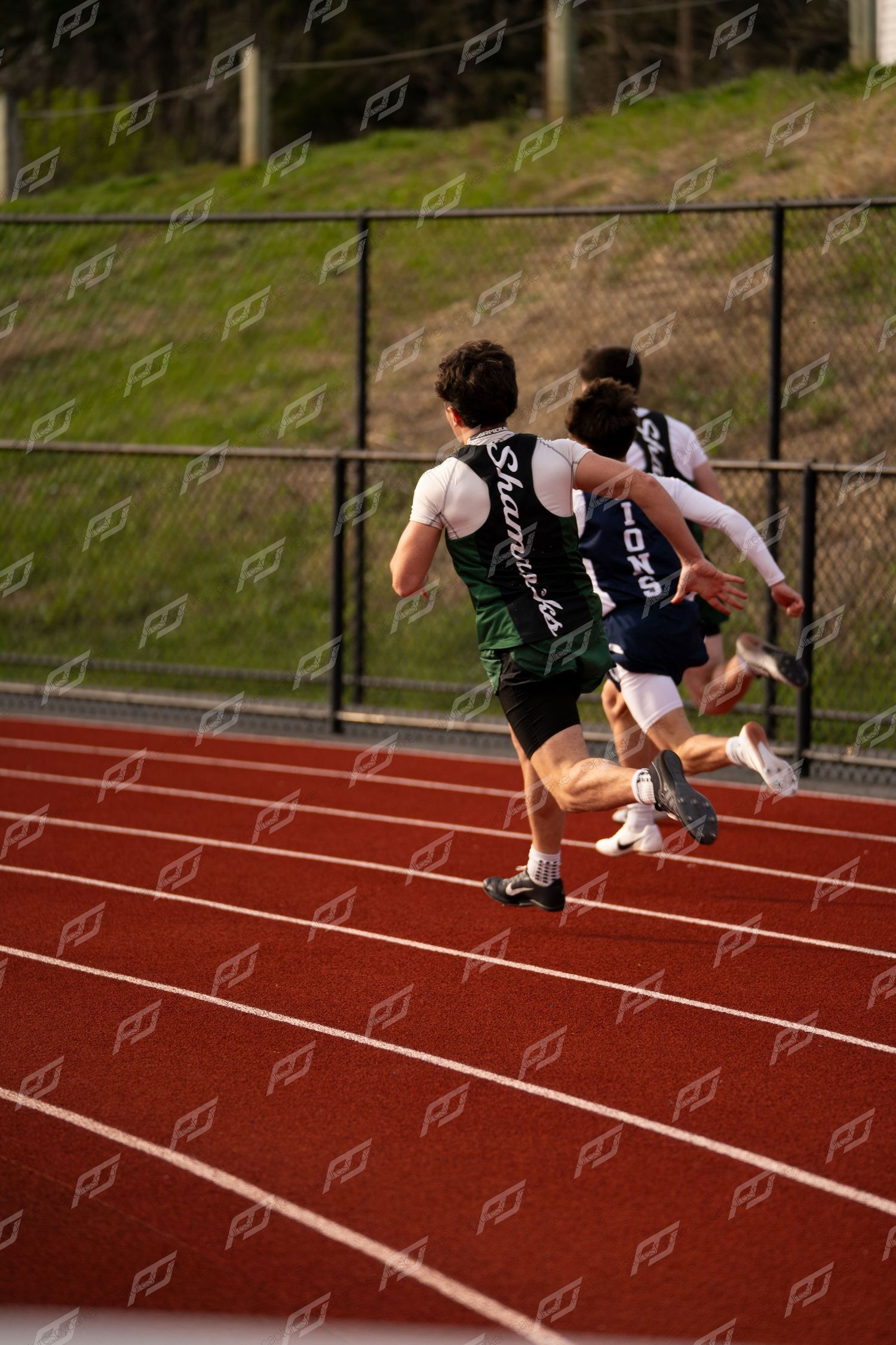 Three high school male sprinters race down red track during competitive track and field meet.