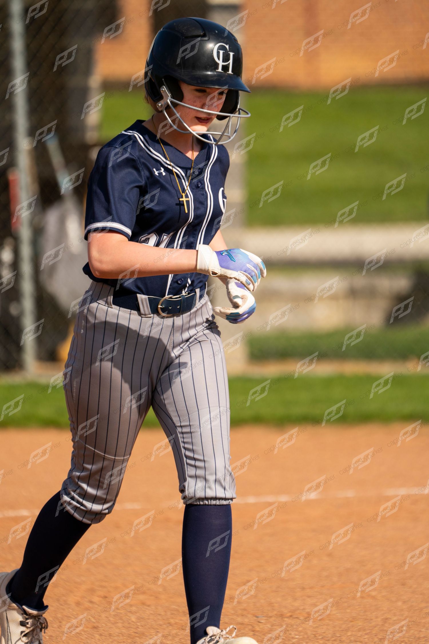 Female softball player in navy CH uniform walks on dirt field holding batting gloves after at-bat.