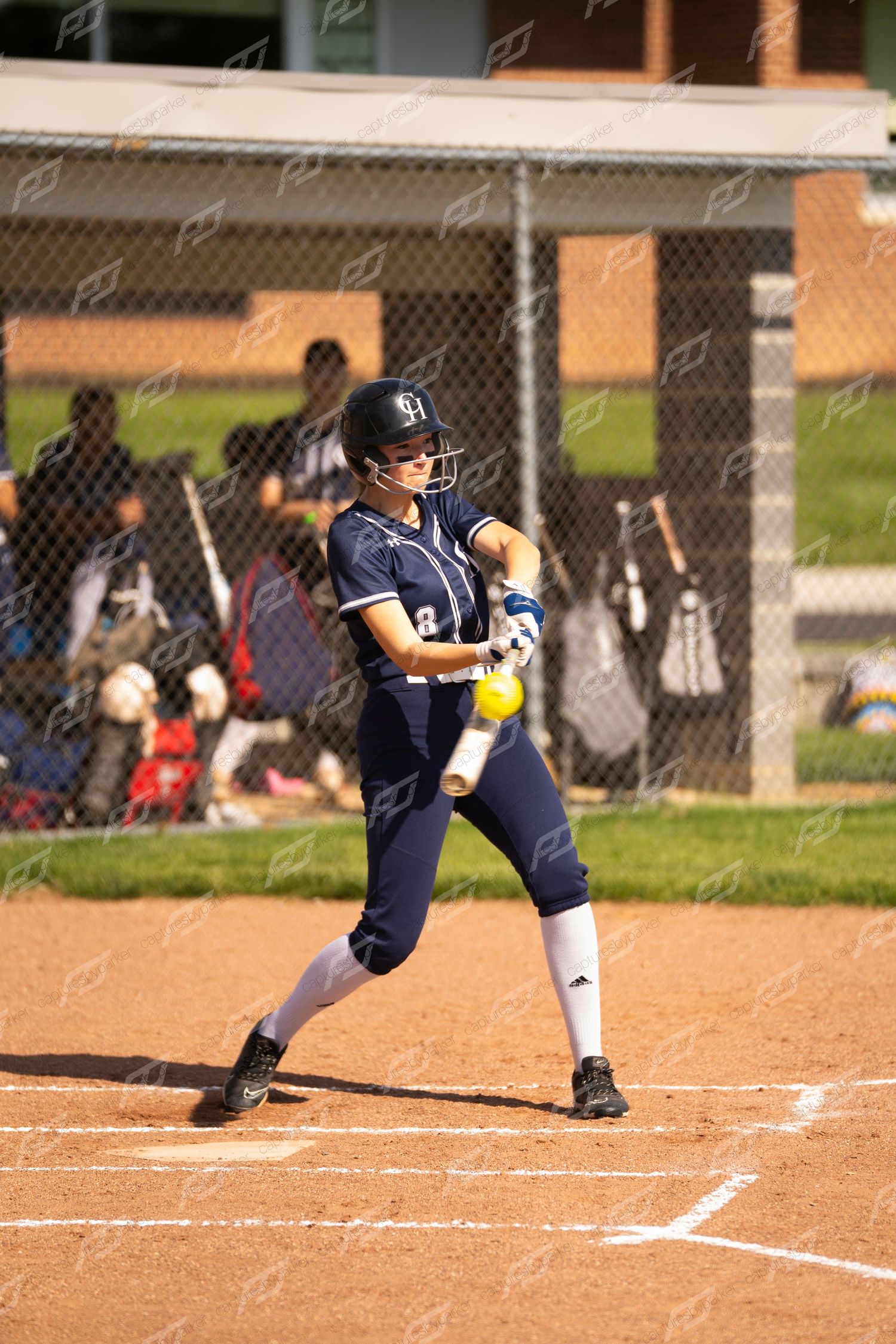 Female softball batter in navy uniform makes contact with yellow ball at home plate.