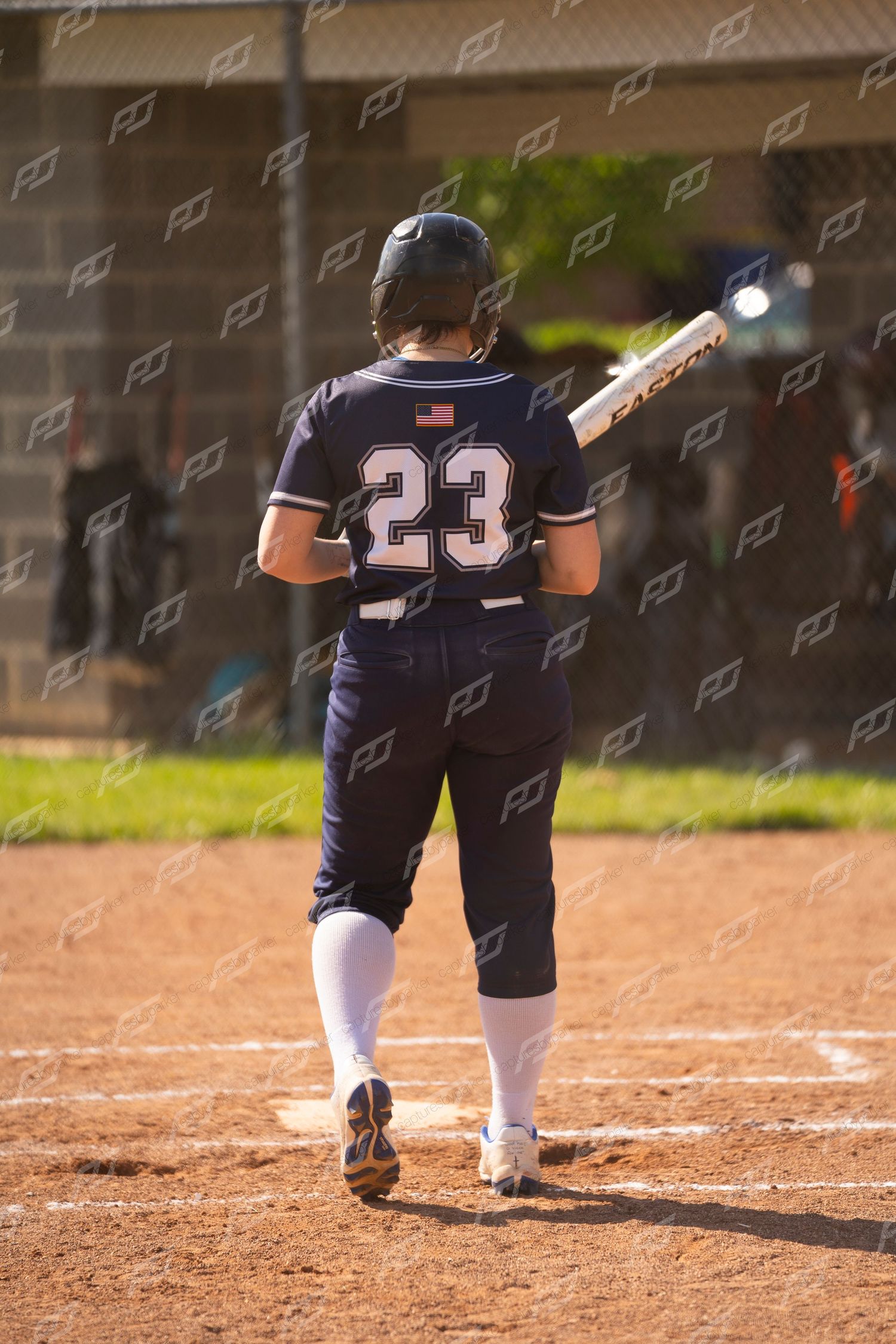 Softball player number 23 in navy uniform walks to the plate carrying a bat.