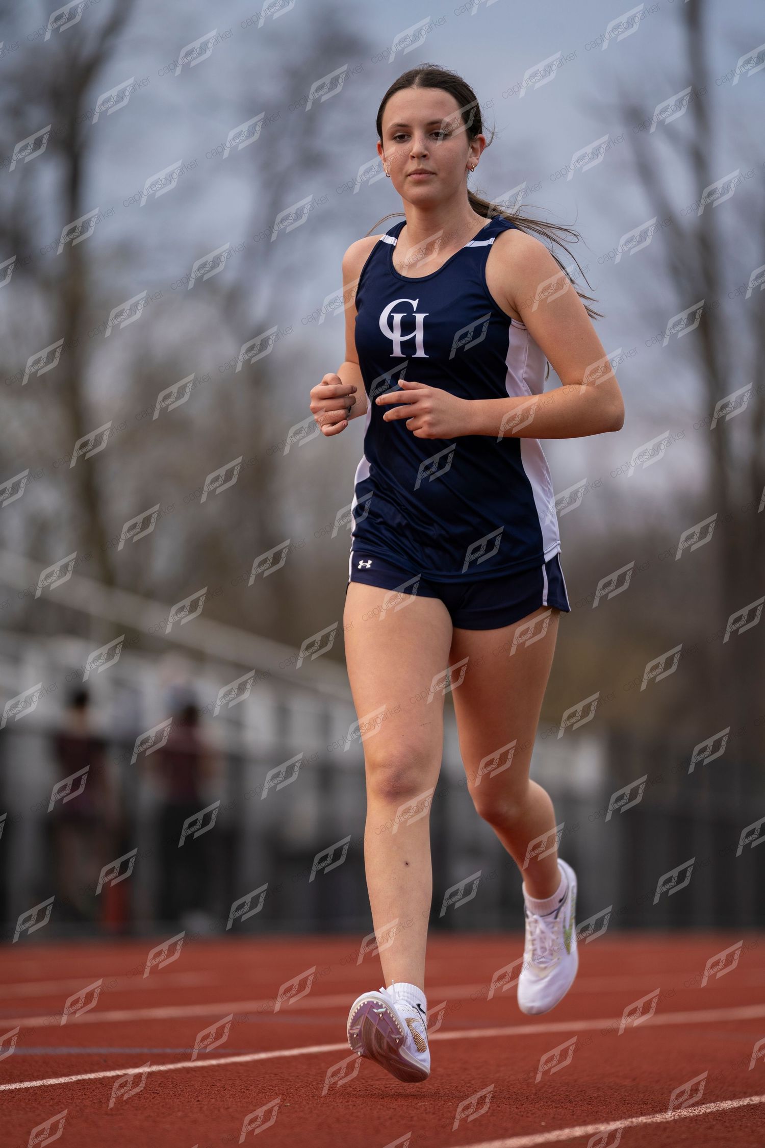 Female track athlete in navy CH uniform running competitively on outdoor red track.