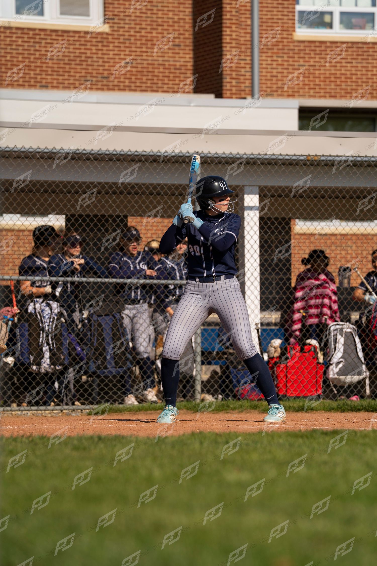 Softball player in navy pinstripe uniform stands ready at home plate gripping a blue bat during an outdoor game.