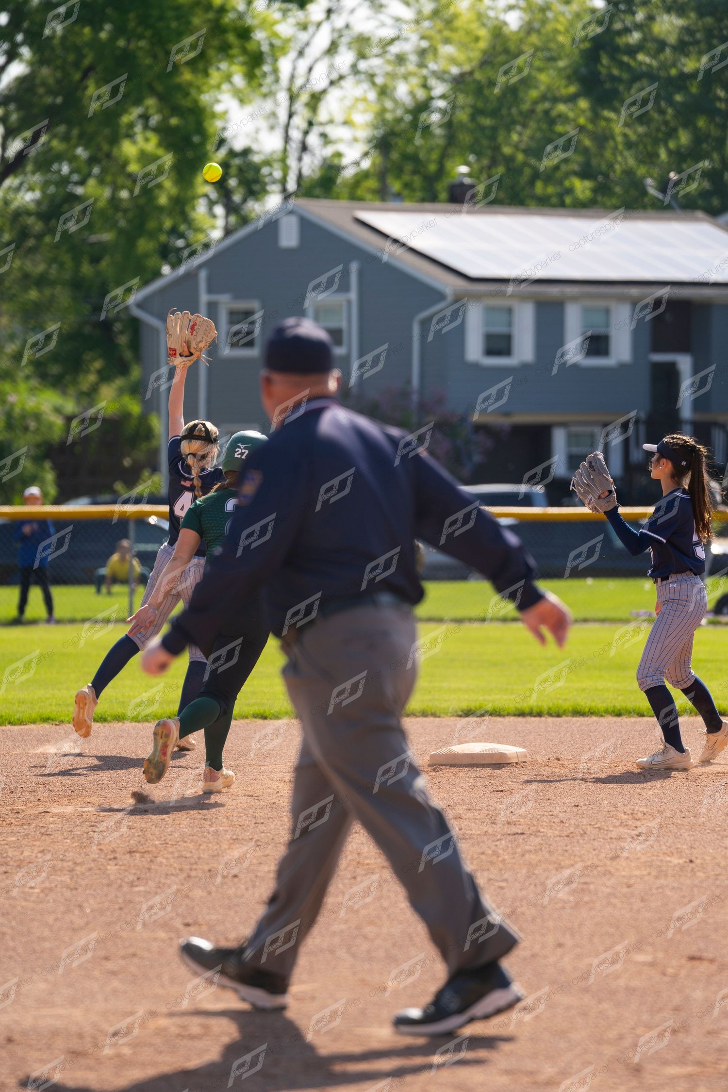 Female softball shortstop in green uniform throws ball as umpire and fielder watch nearby.