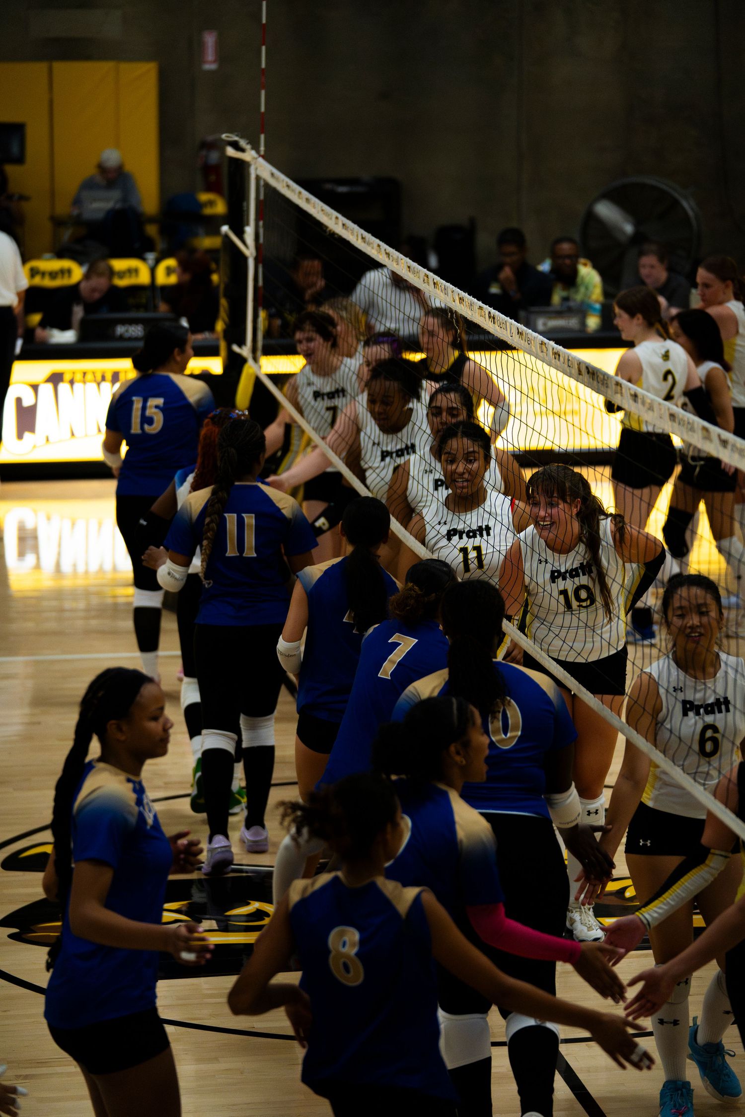 Two volleyball teams shake hands at the net after a match, wearing blue and white uniforms on an indoor court.