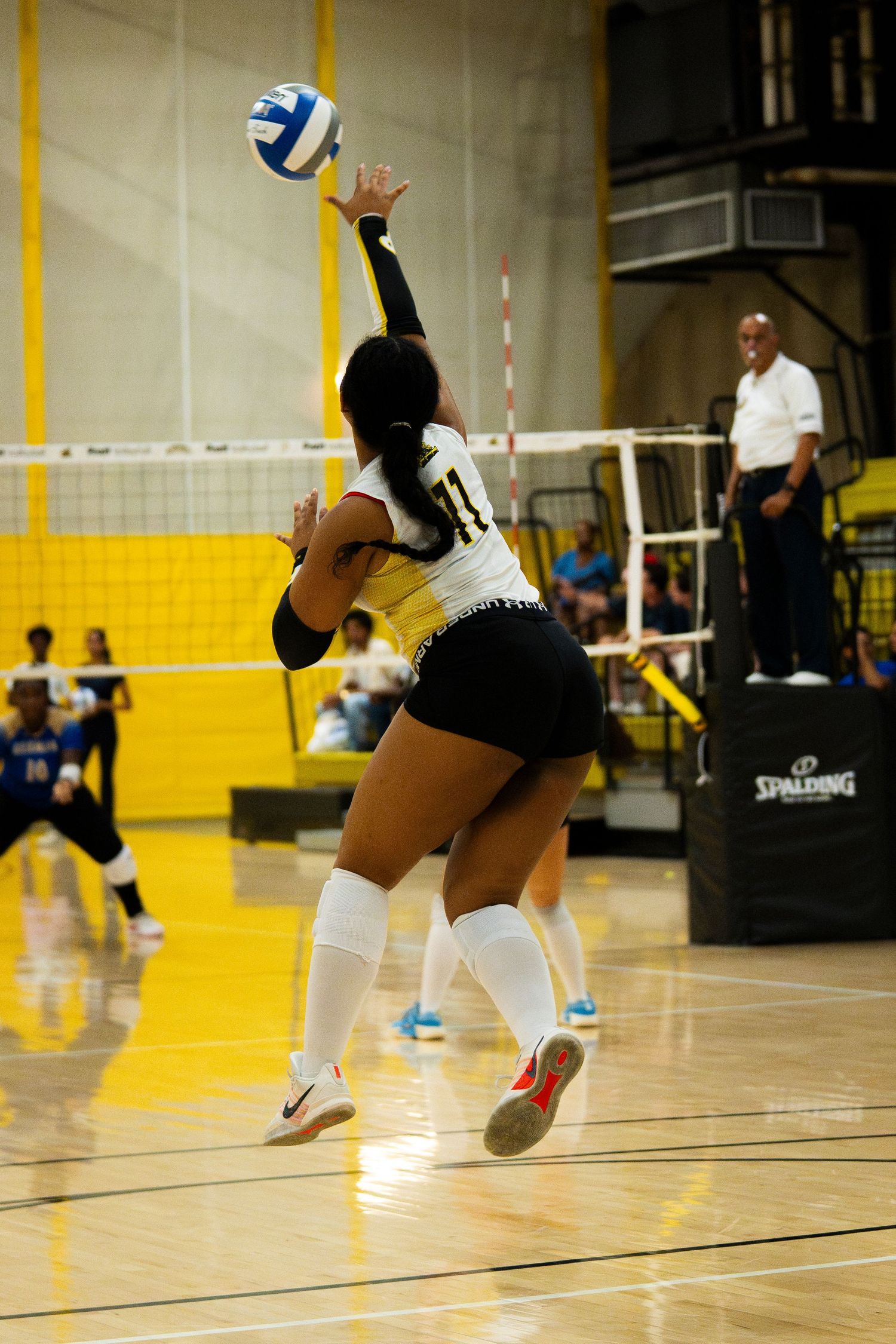 Female volleyball player in yellow uniform jumping to spike the ball during an indoor match on a hardwood court.