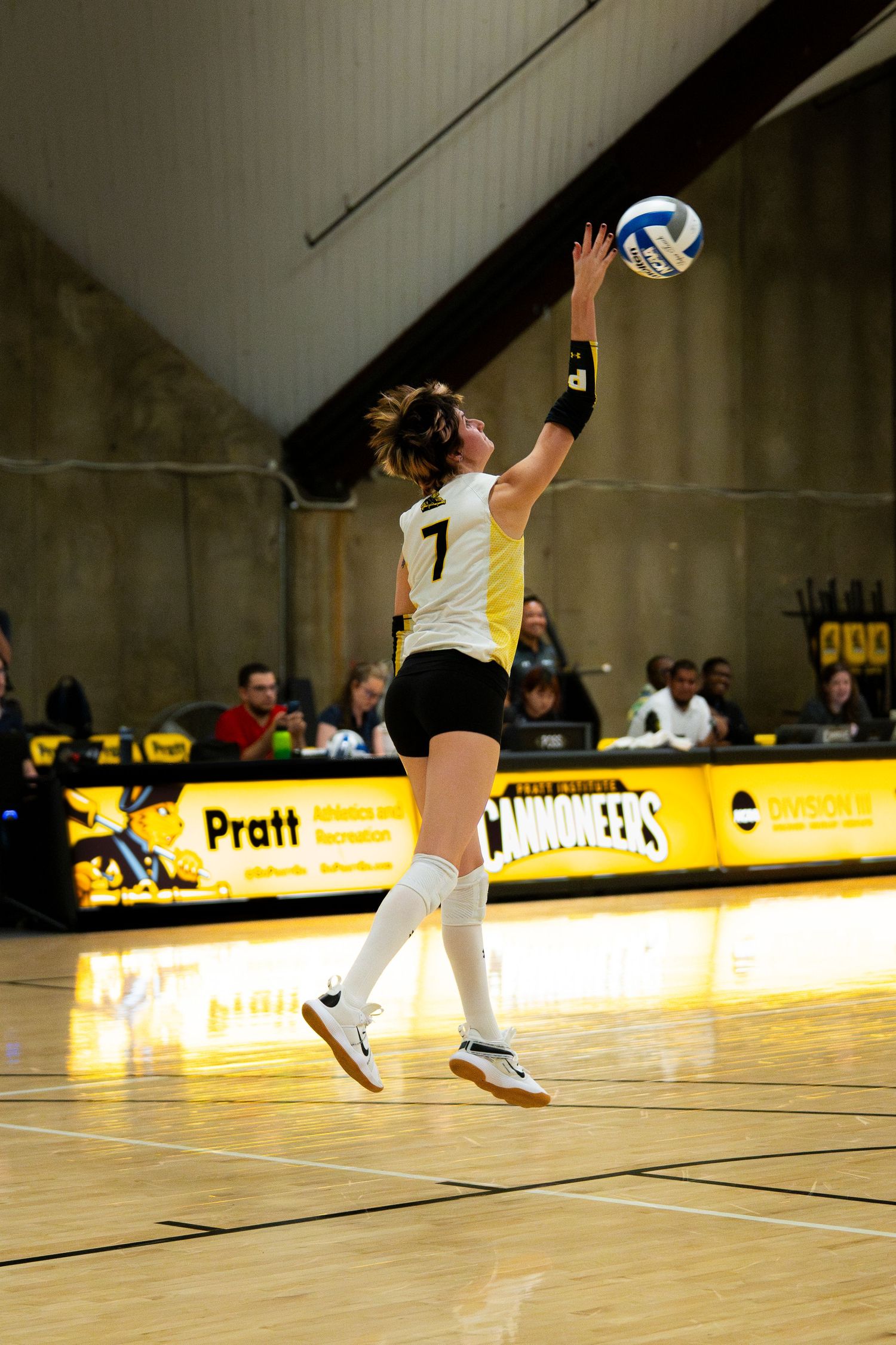 Female volleyball player #7 in black and white uniform serves the ball during an indoor match at Pratt Cannoneer gymnasium.
