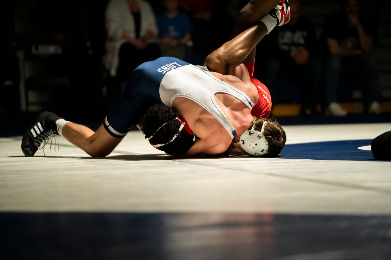 Two wrestlers grapple on the mat, one in blue pinning the other in red and white during a wrestling match.